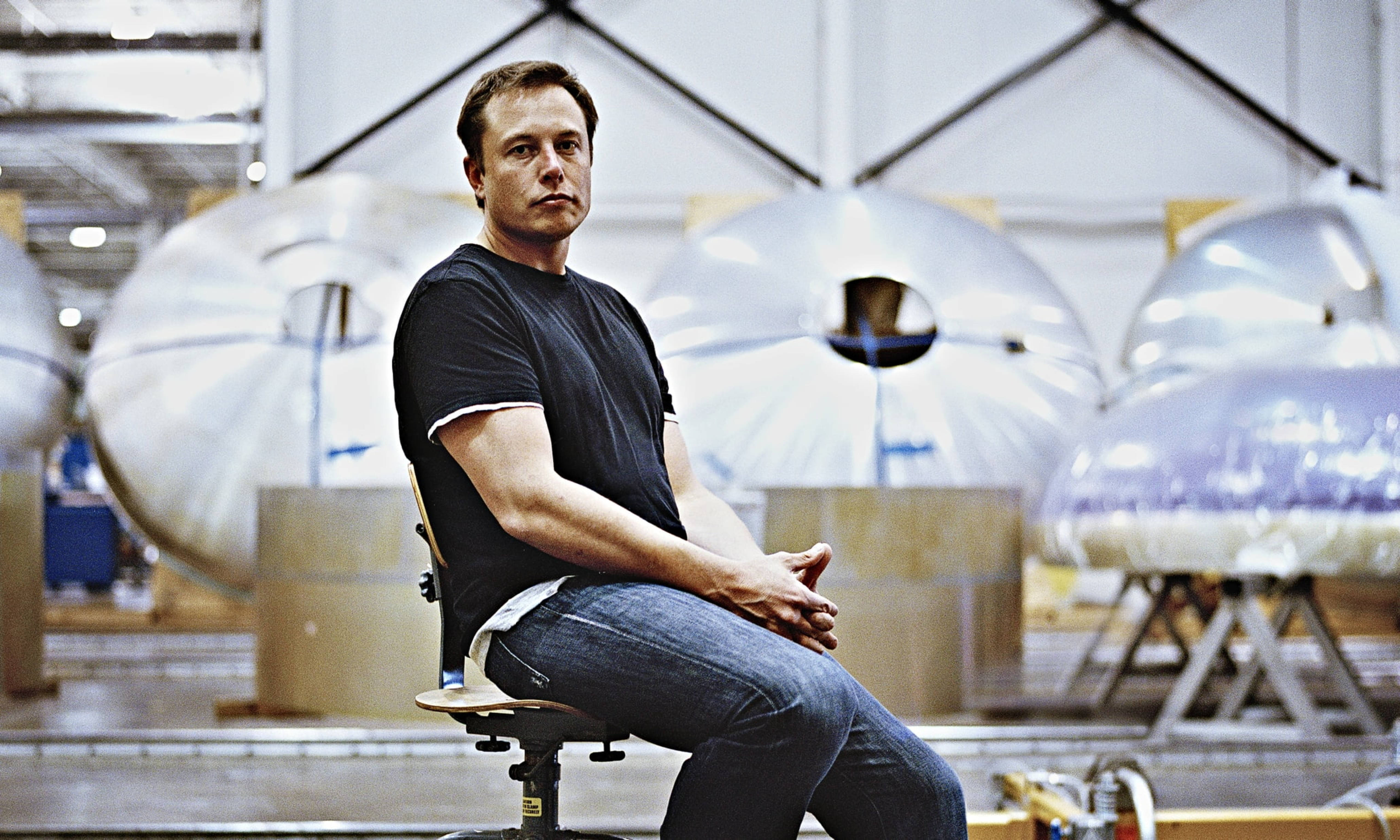 Elon 桌布 壁紙 — 伊隆·馬斯克坐在工廠的凳子上，周圍是大型銀色火箭引擎，在明亮的工業照明下。