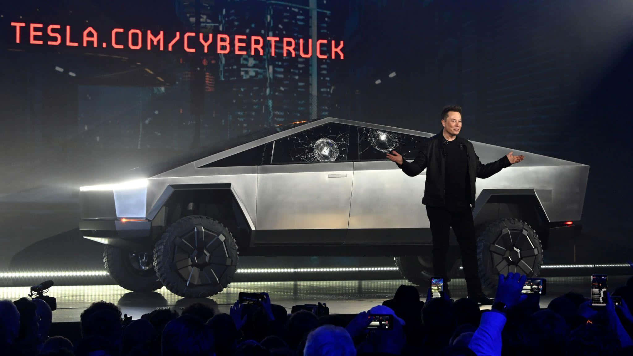 Tesla Cybertruck 桌布 壁紙 — 伊隆·馬斯克站在舞台上，在明亮的舞台燈光下，在一輛車窗破碎的銀色 Tesla Cybertruck 旁邊做手勢。