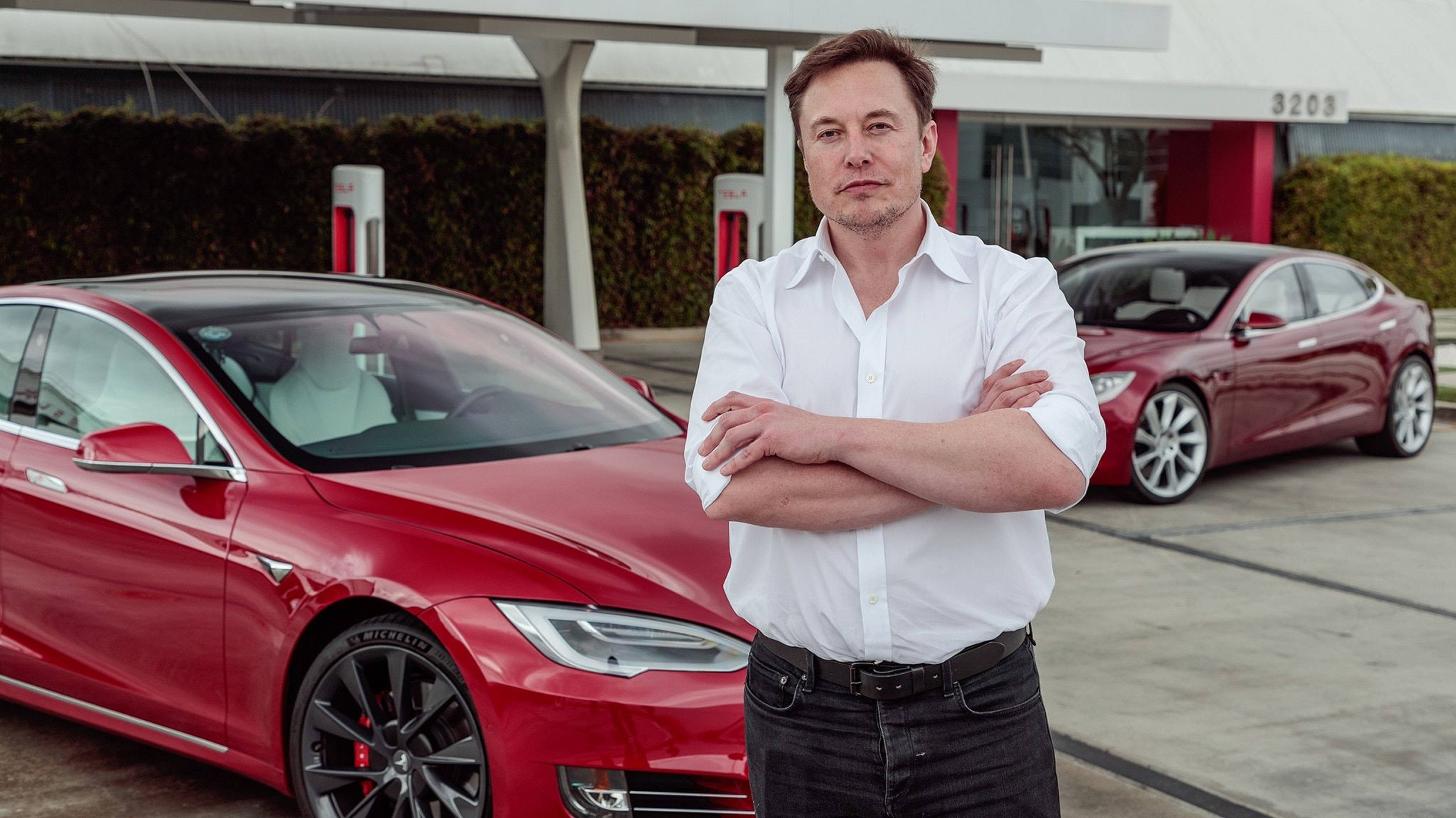 Elon 桌布 壁紙 — 伊隆·馬斯克自信地交叉雙臂站在充電站旁的一輛紅色特斯拉汽車旁邊。