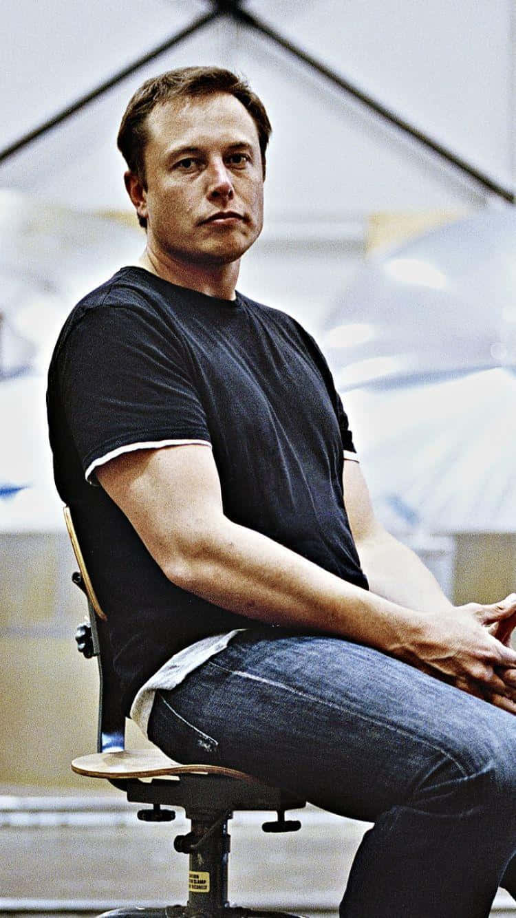 伊隆·馬斯克 桌布 壁紙 — 伊隆·馬斯克隨意地坐在明亮工業空間的凳子上，表情專注。