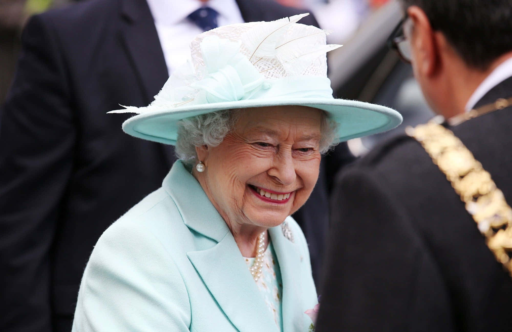 Queen Elizabeth 桌布 壁紙 — 伊麗莎白二世女王在公開活動中身穿淺藍色套裝和羽毛帽，溫暖地綻放笑容。