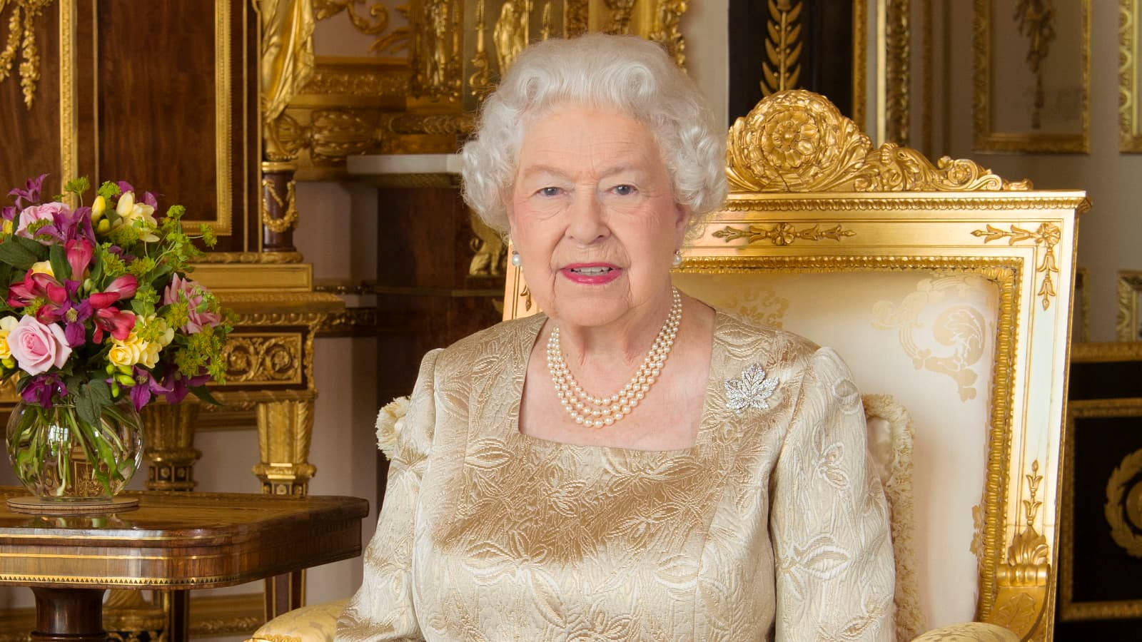 Queen Elizabeth 桌布 壁紙 — 伊麗莎白二世女王威嚴地坐在一個鍍金的寶座廳中，廳內裝飾著華麗的金色家具和鮮豔的花藝佈置。