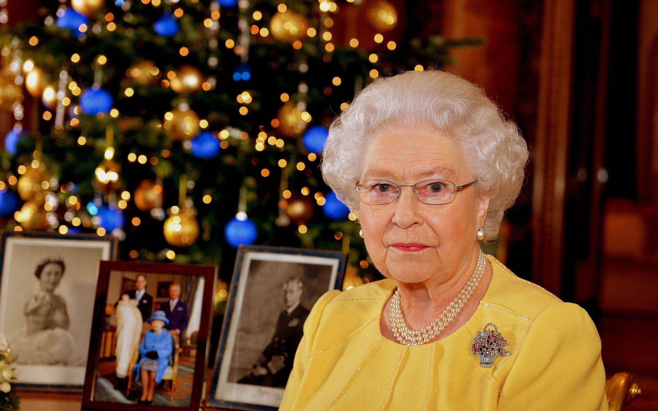 Queen Elizabeth 桌布 壁紙 — 伊麗莎白二世女王身穿黃色服裝，坐在一棵裝飾著發光飾物和裱框家庭照片的節日聖誕樹前。