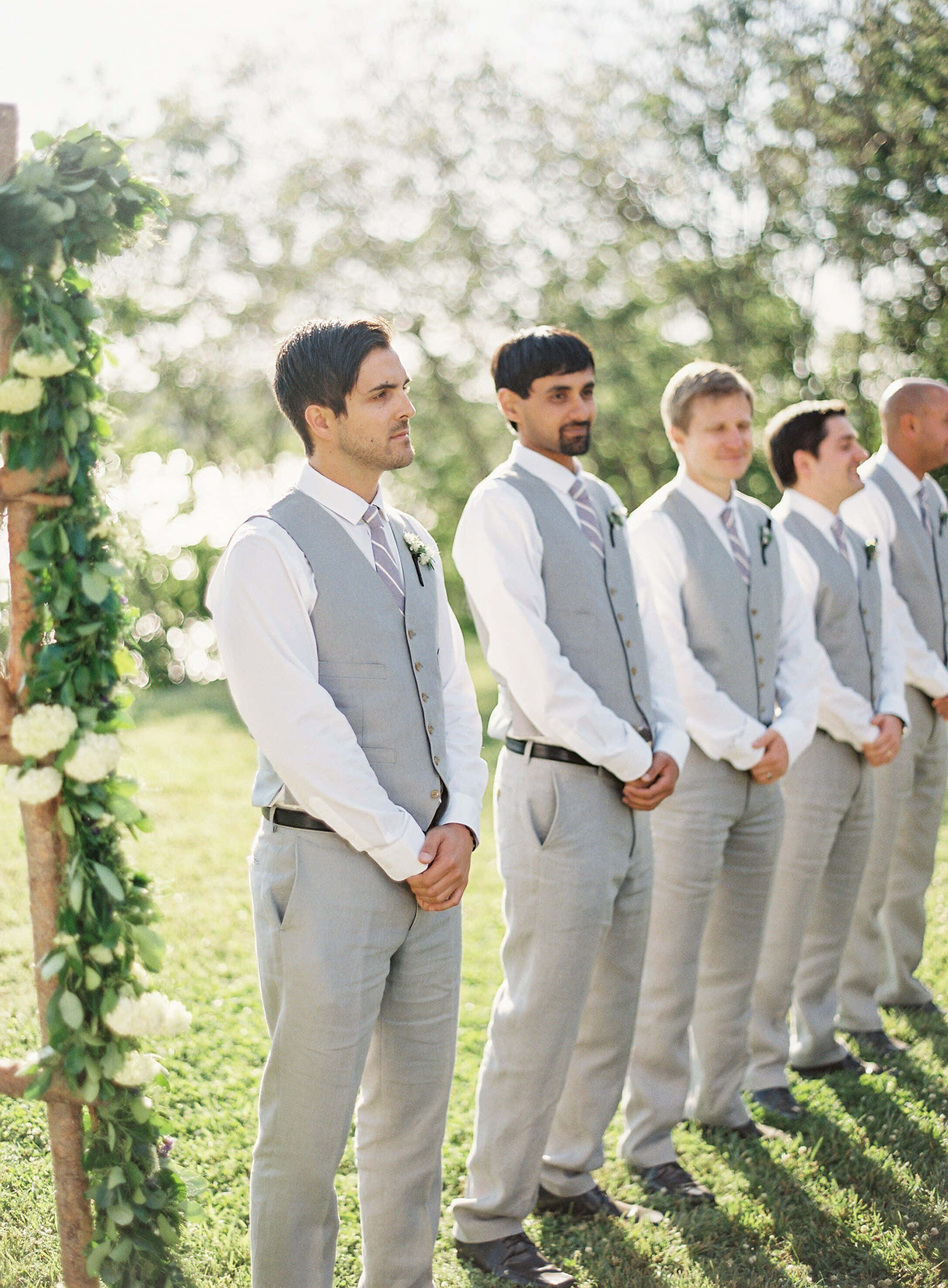 Groomsman 桌布 壁紙 — 伴郎們穿著灰色背心和白色襯衫，在明亮的陽光下站在草地上，旁邊有一個花拱門。