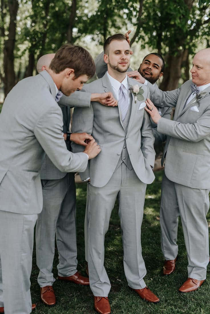 Groomsmen 桌布 壁紙 — 伴郎們穿著相配的灰色西裝，在陽光明媚的森林空地中調整新郎的翻領和領帶。