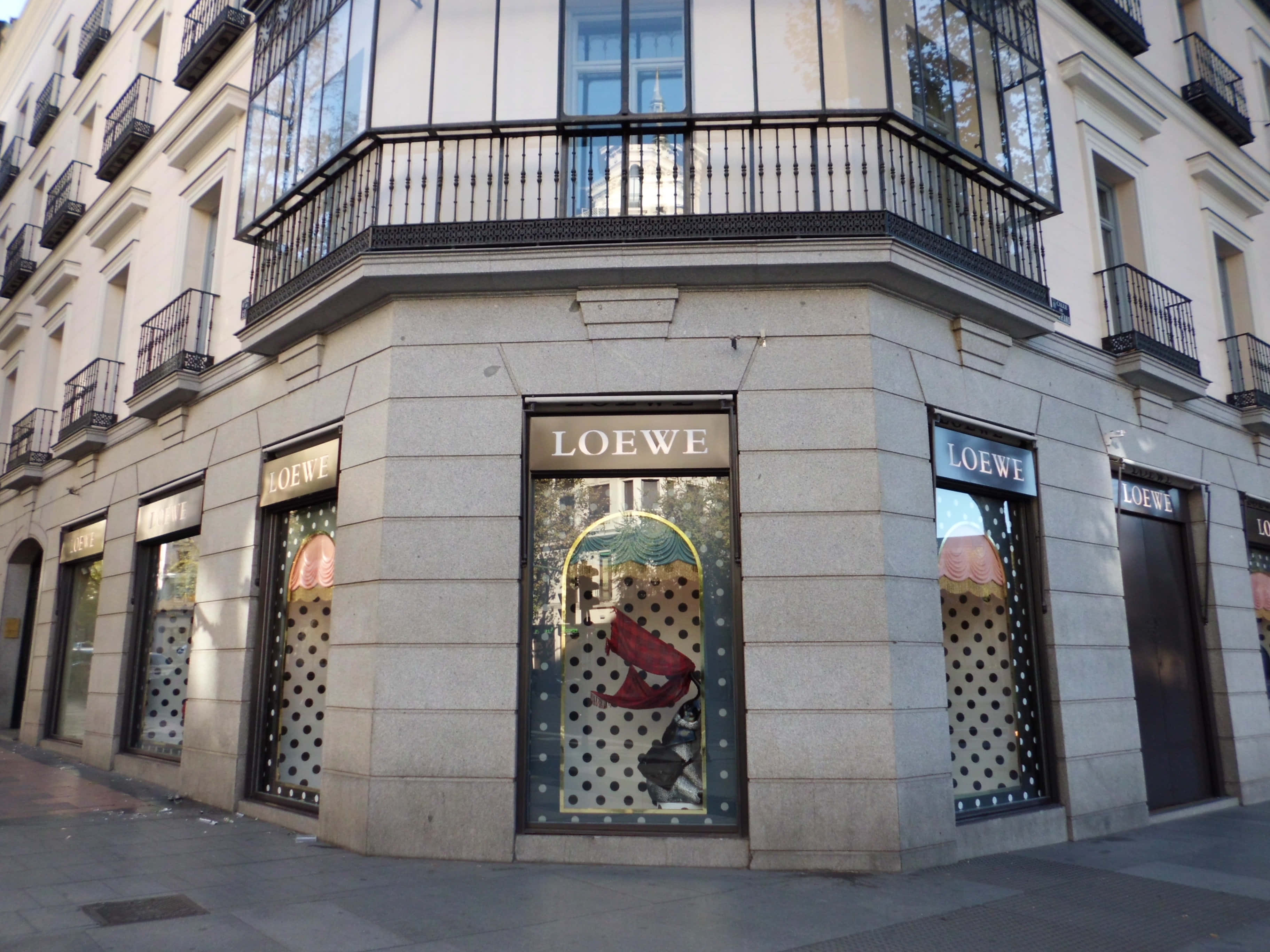 Building 桌布 壁紙 — 位於角落的Loewe精品店，飾有圓點櫥窗展示和黑色鐵藝陽台，襯托米色石牆。