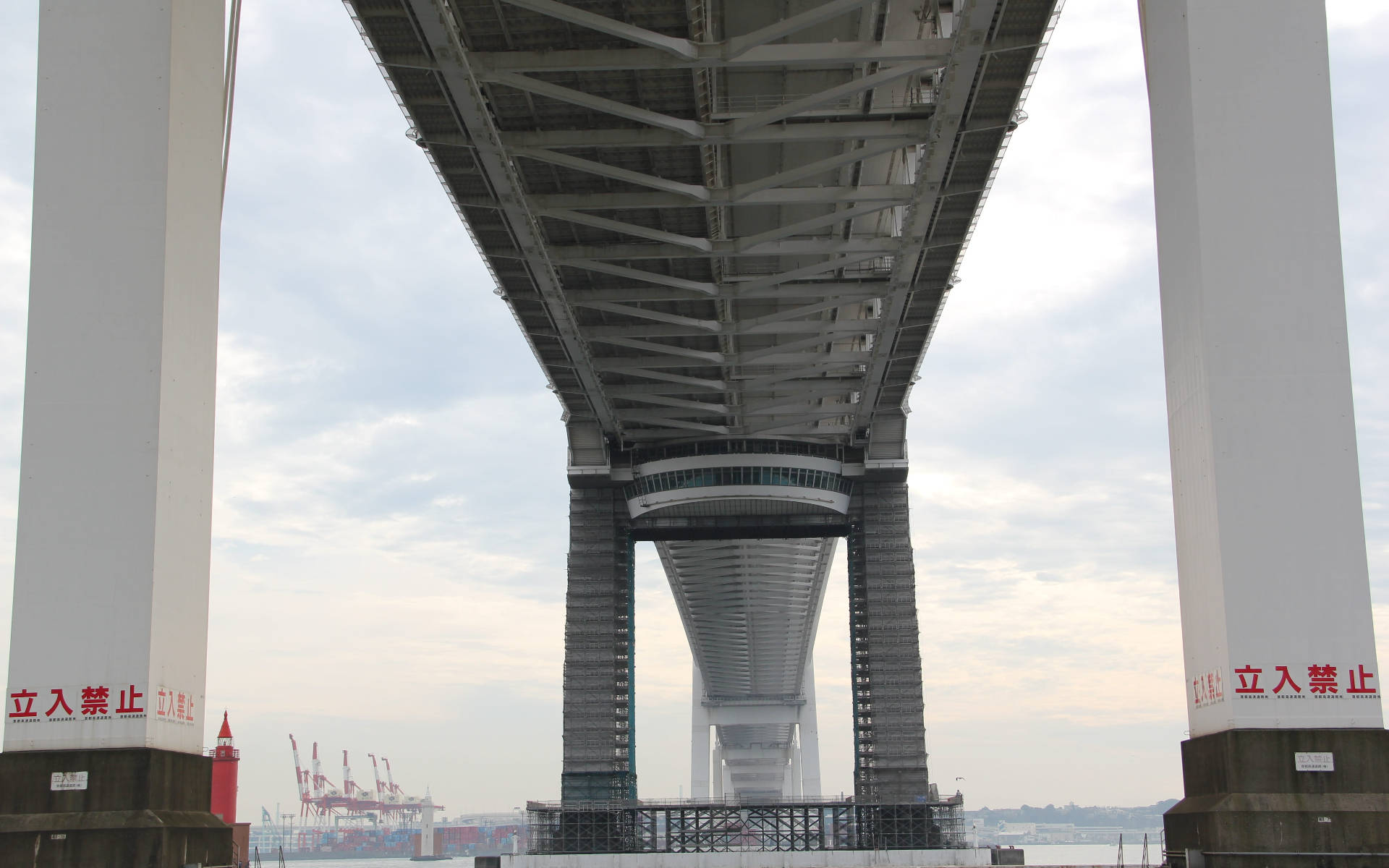 Yokohama 桌布 壁紙 — 低角度向上觀看現代吊橋的巨大鋼筋混凝土結構，遠處可見工業港口起重機，天空多雲。
