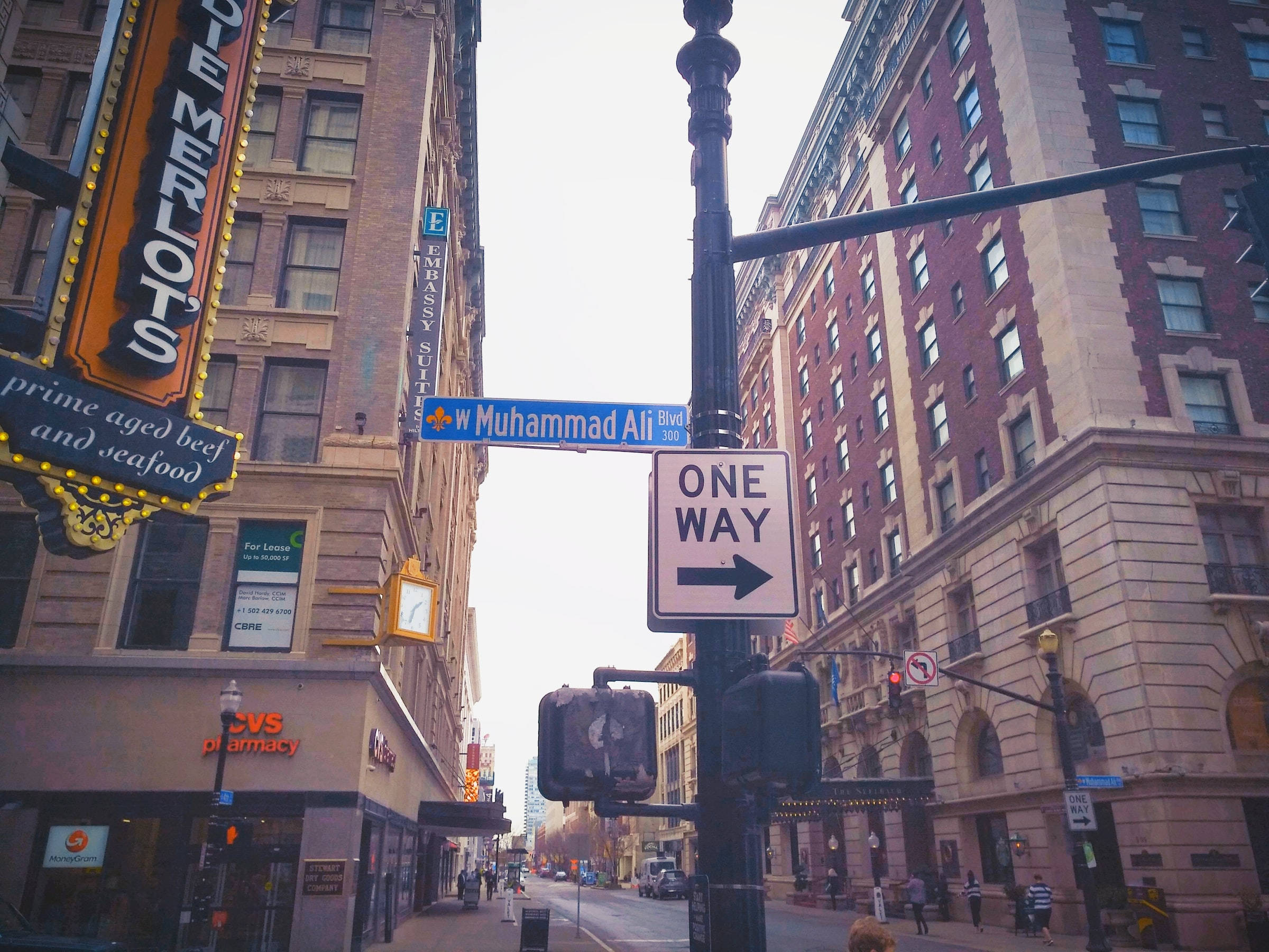 城市 桌布 壁紙 — 低角度城市街景，由高樓大廈框住，前景有「Muhammad Ali Blvd」標誌和「ONE WAY」街道標誌。