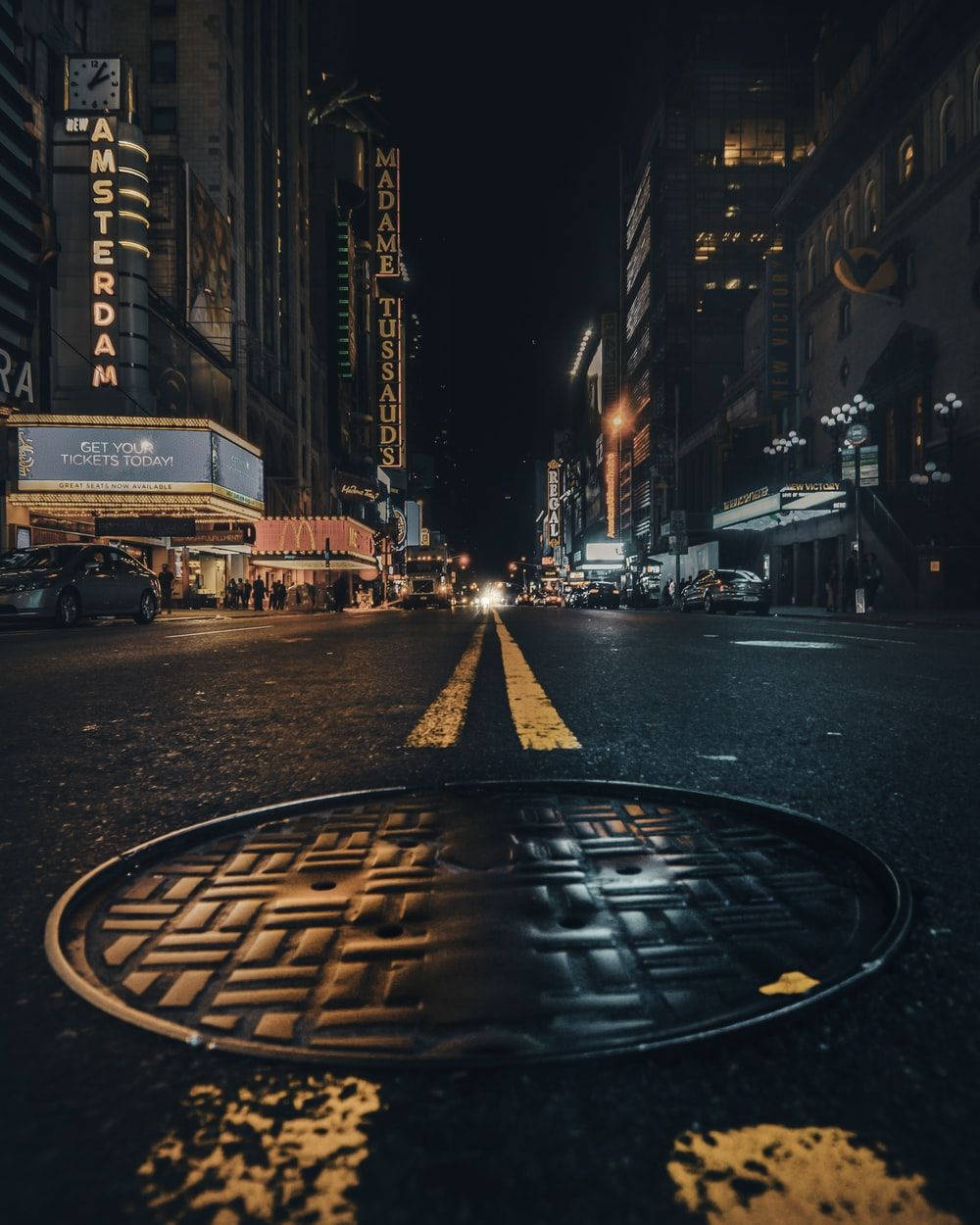 City Street 桌布 壁紙 — 低角度夜拍濕漉漉的城市街道，前景有沙井蓋，以及閃爍的 Madame Tussauds 和 Amsterdam 劇院霓虹招牌。