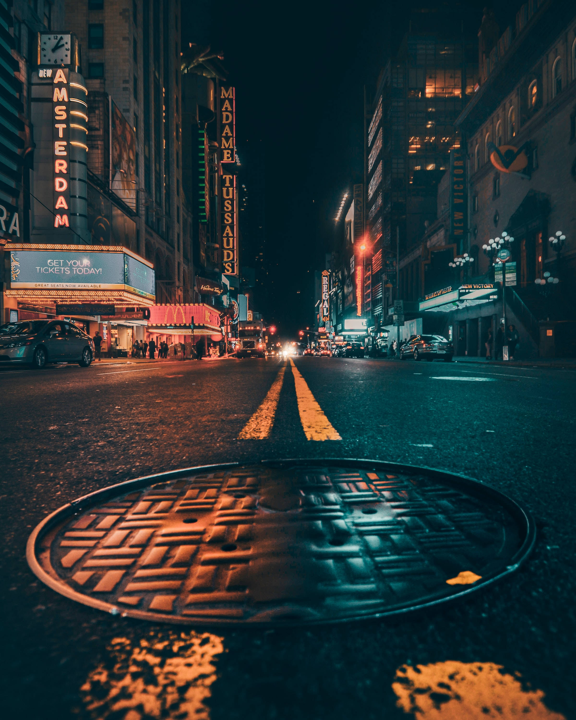 夜空 桌布 壁紙 — 低角度夜拍濕漉漉的城市街道，前景有沙井蓋，背景有杜莎夫人蠟像館和阿姆斯特丹劇院的發光霓虹招牌。