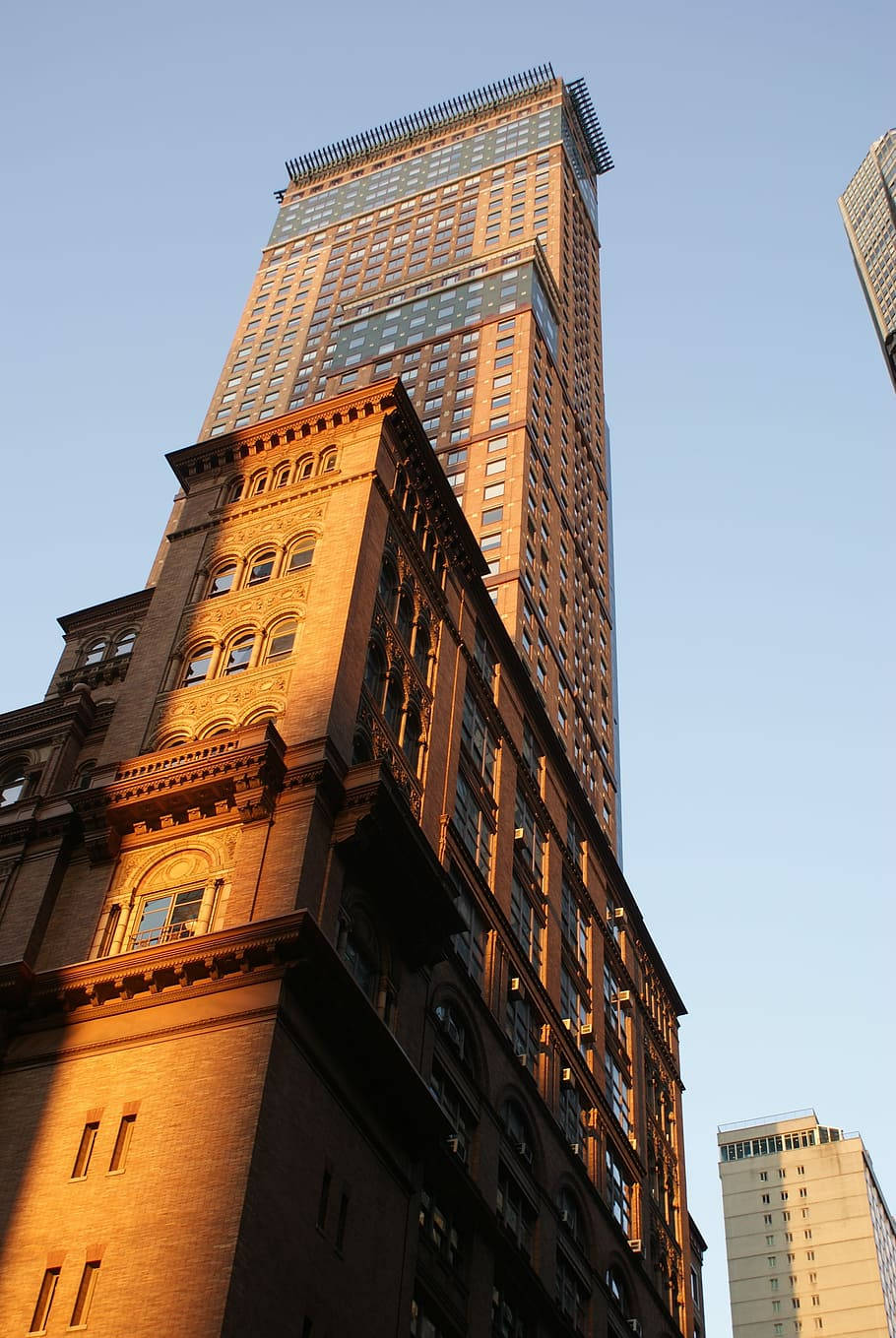建築 桌布 壁紙 — 低角度拍攝一座歷史悠久的褐石建築，沐浴在金色的日落光芒中，背景是清澈的藍天，一座現代摩天大樓聳立其後。