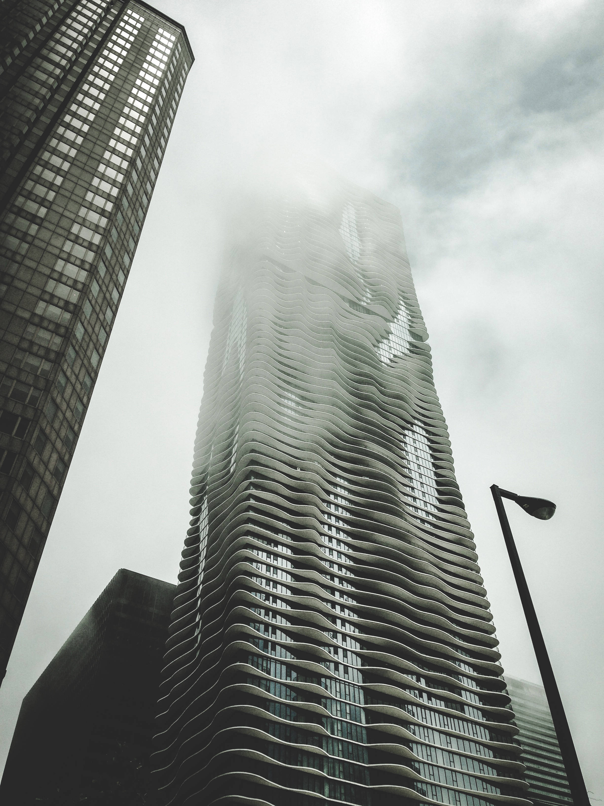 Futuristic City 桌布 壁紙 — 低角度拍攝一座被薄霧籠罩的現代摩天大樓，帶有波浪形陽台，聳立在城市街道上，天空多雲。