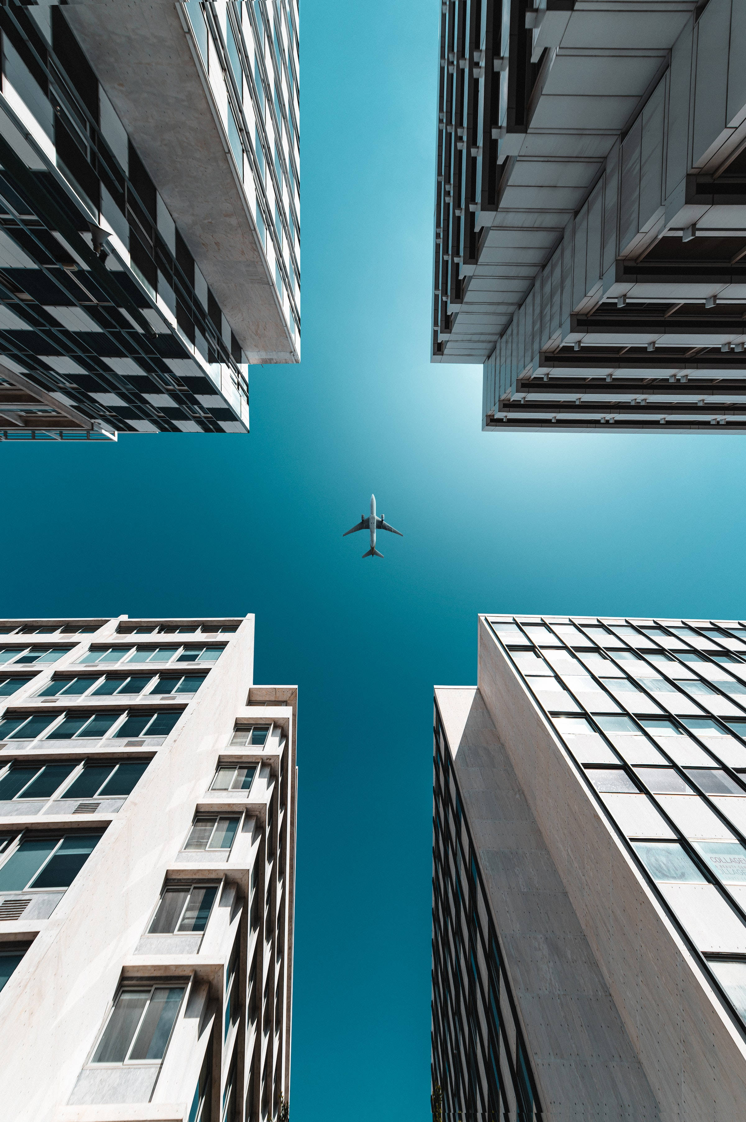 Skyscraper 桌布 壁紙 — 低角度拍攝一架銀色飛機飛過晴朗藍天，以高聳的現代建築為框。