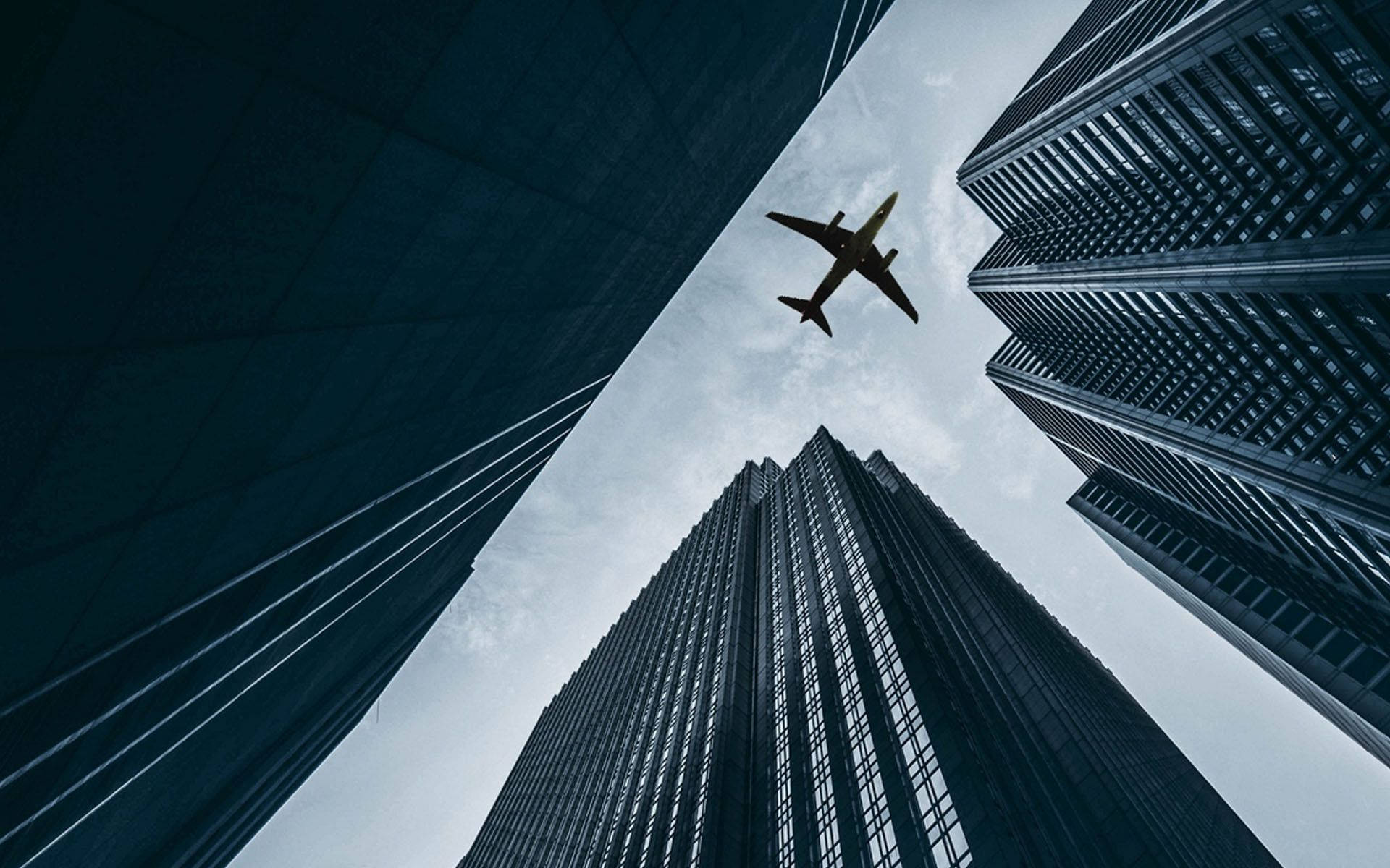  壁紙 — 低角度拍攝一架飛機在陰天天空下飛越摩天大樓之間。