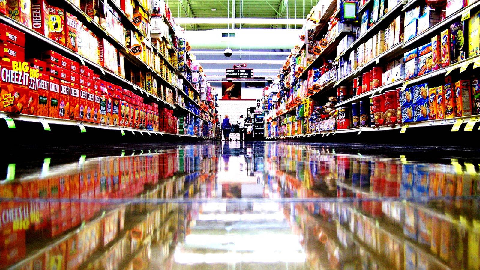 Shopping 桌布 壁紙 — 低角度拍攝一條光線明亮的超市通道，反射地板映照出色彩繽紛的零食盒。