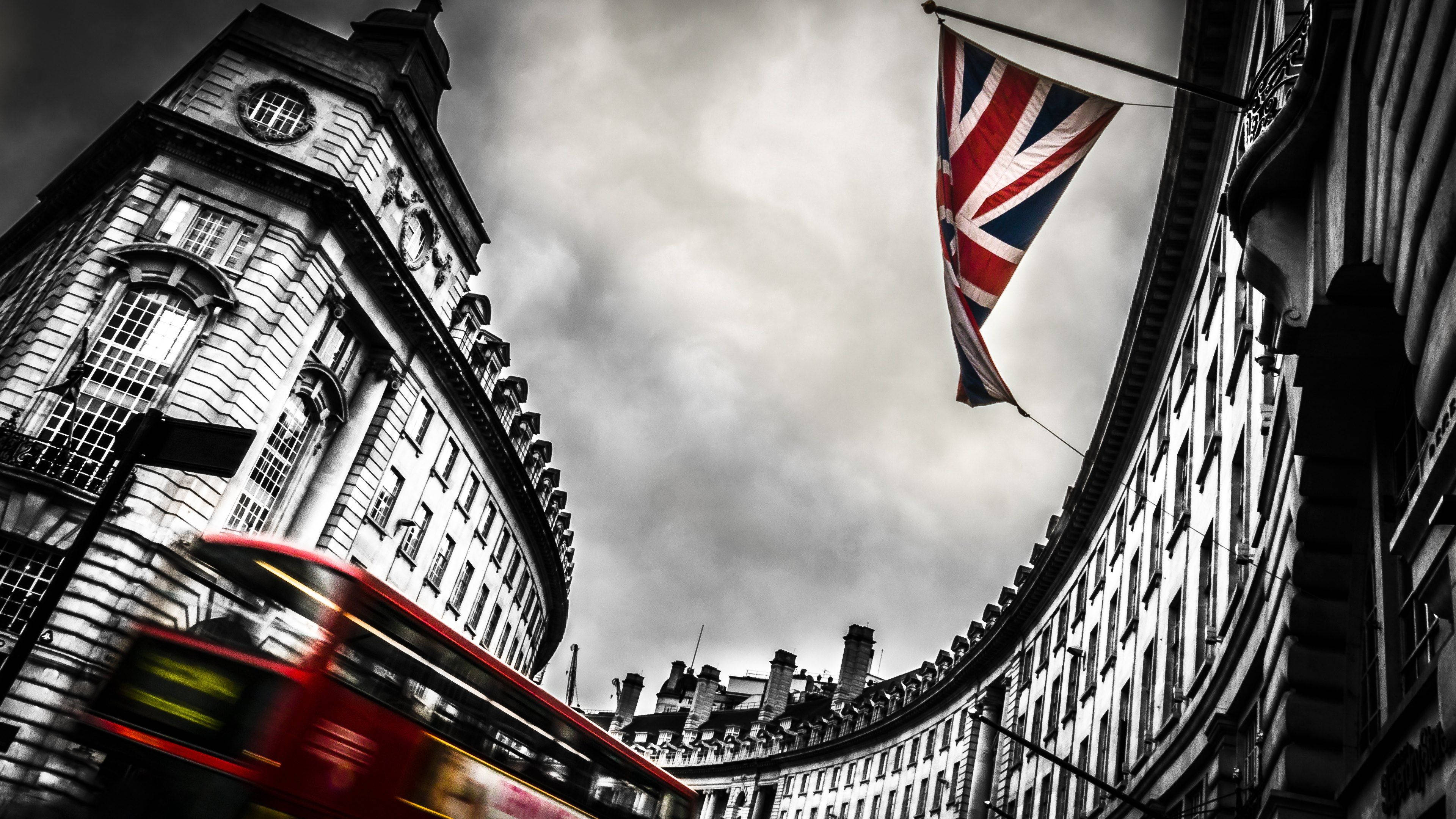 United Kingdom Flag 桌布 壁紙 — 低角度拍攝一輛紅色雙層巴士在陰沉天空下，帶有飄揚的英國國旗，飛馳經過歷史悠久的倫敦建築。