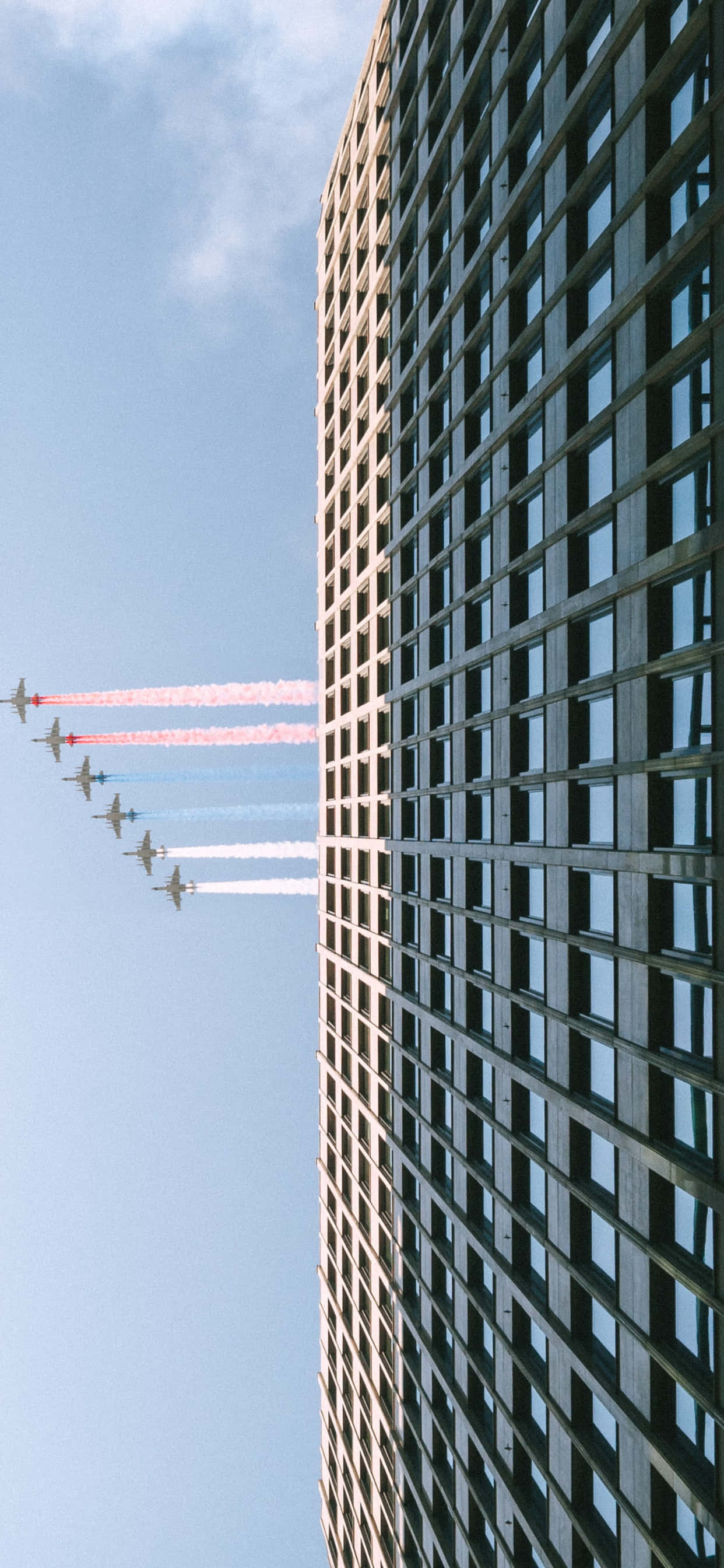 iPhone Xs Max Small Planes 桌布 壁紙 — 低角度拍攝戰鬥機編隊飛行，拖曳著彩色煙霧，掠過高聳的摩天大樓，背景是清澈的藍天。