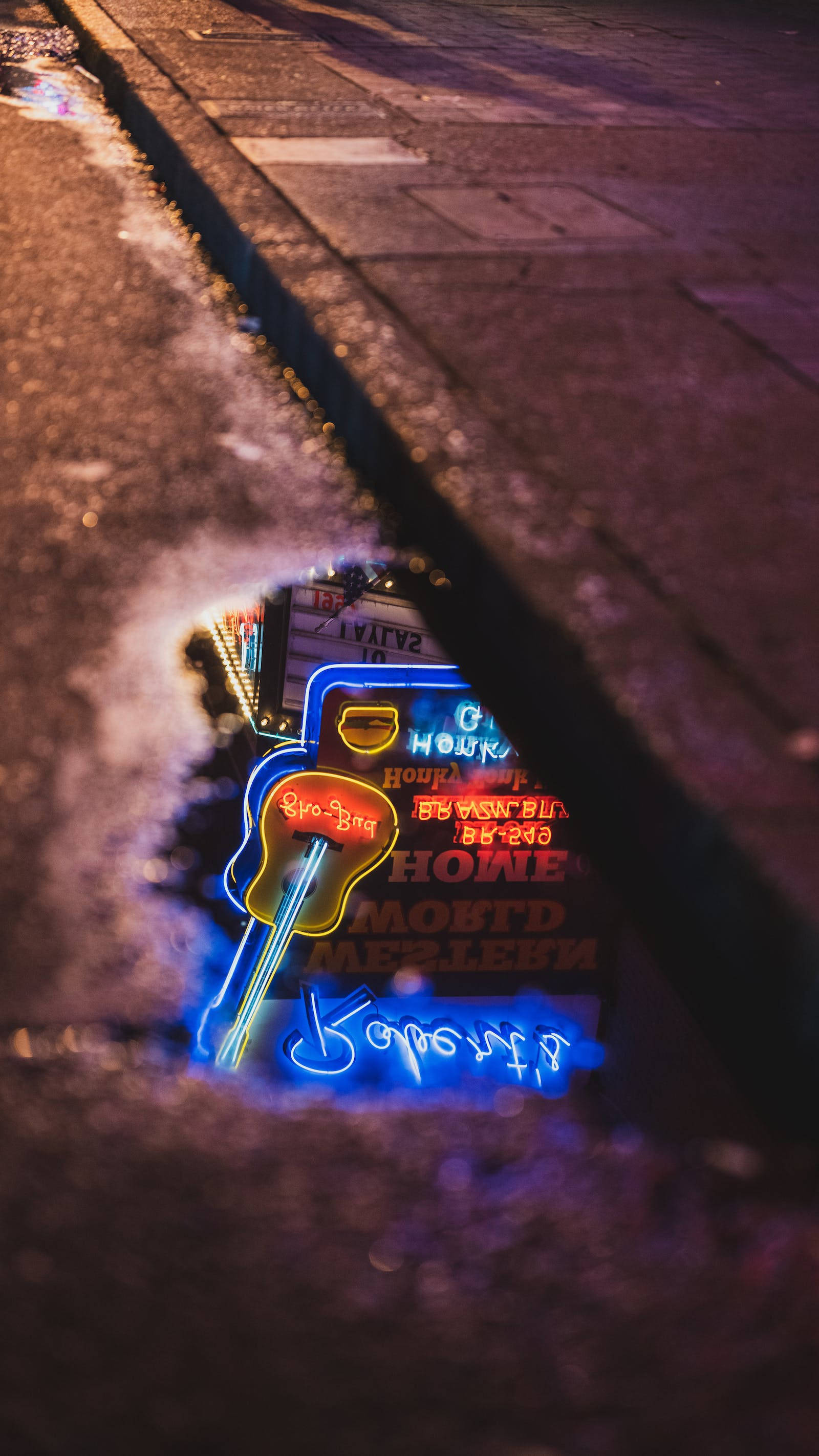 Neon Phone 桌布 壁紙 — 低角度拍攝捕捉到一個充滿活力的吉他形狀霓虹燈牌，在夜晚透過濕漉漉的城市人行道上的水坑發光。