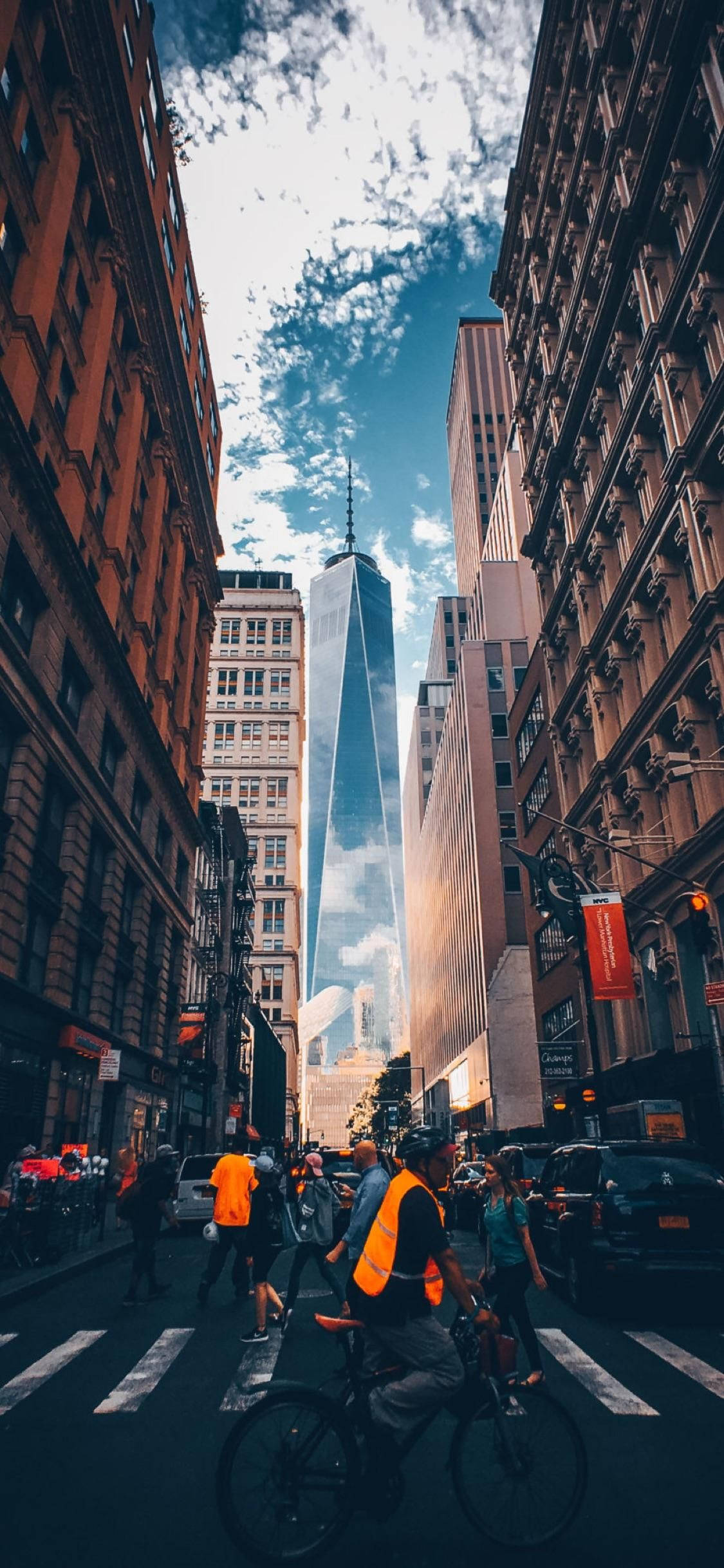 new york city iPhone x 桌布 壁紙 — 低角度拍攝捕捉到一名身穿橙色背心的自行車騎士在紐約市過馬路，自由塔聳立在歷史悠久的褐砂石建築之上，天空部分多雲而湛藍。