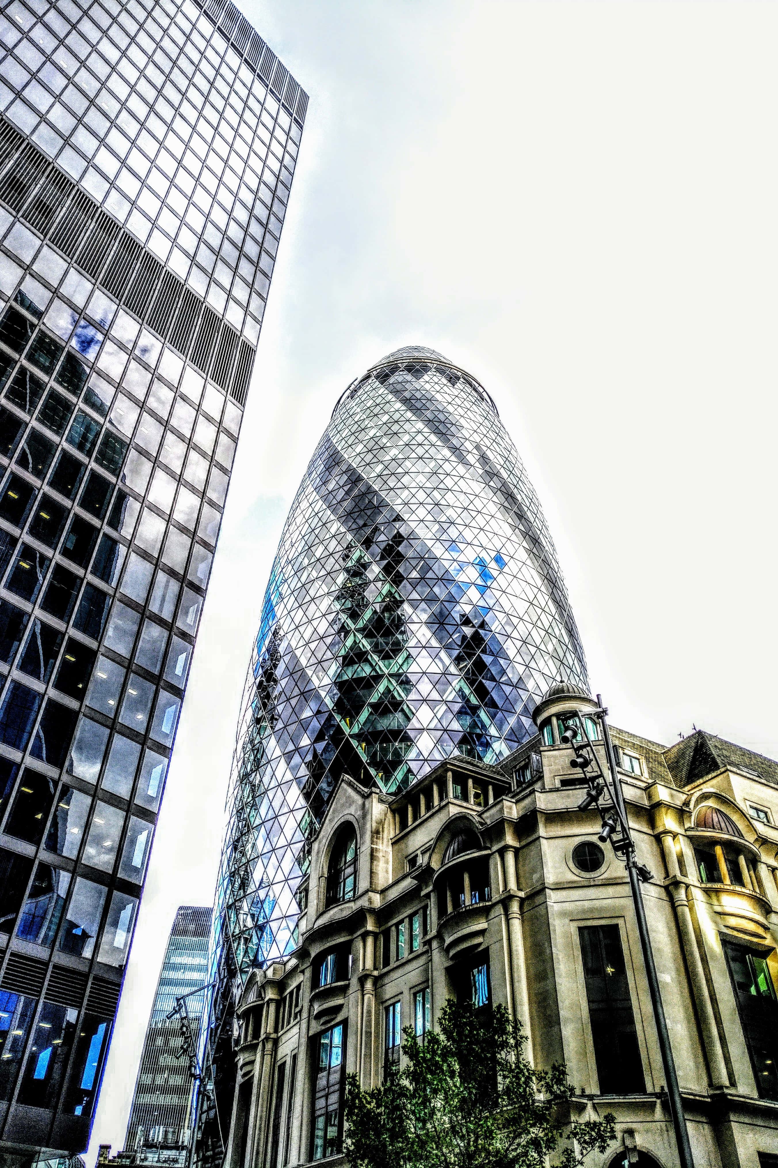 Skyscraper 桌布 壁紙 — 低角度拍攝捕捉到標誌性的Gherkin摩天大樓在晴朗天空下反射天空和雲彩，旁邊是一座歷史悠久的石建築。