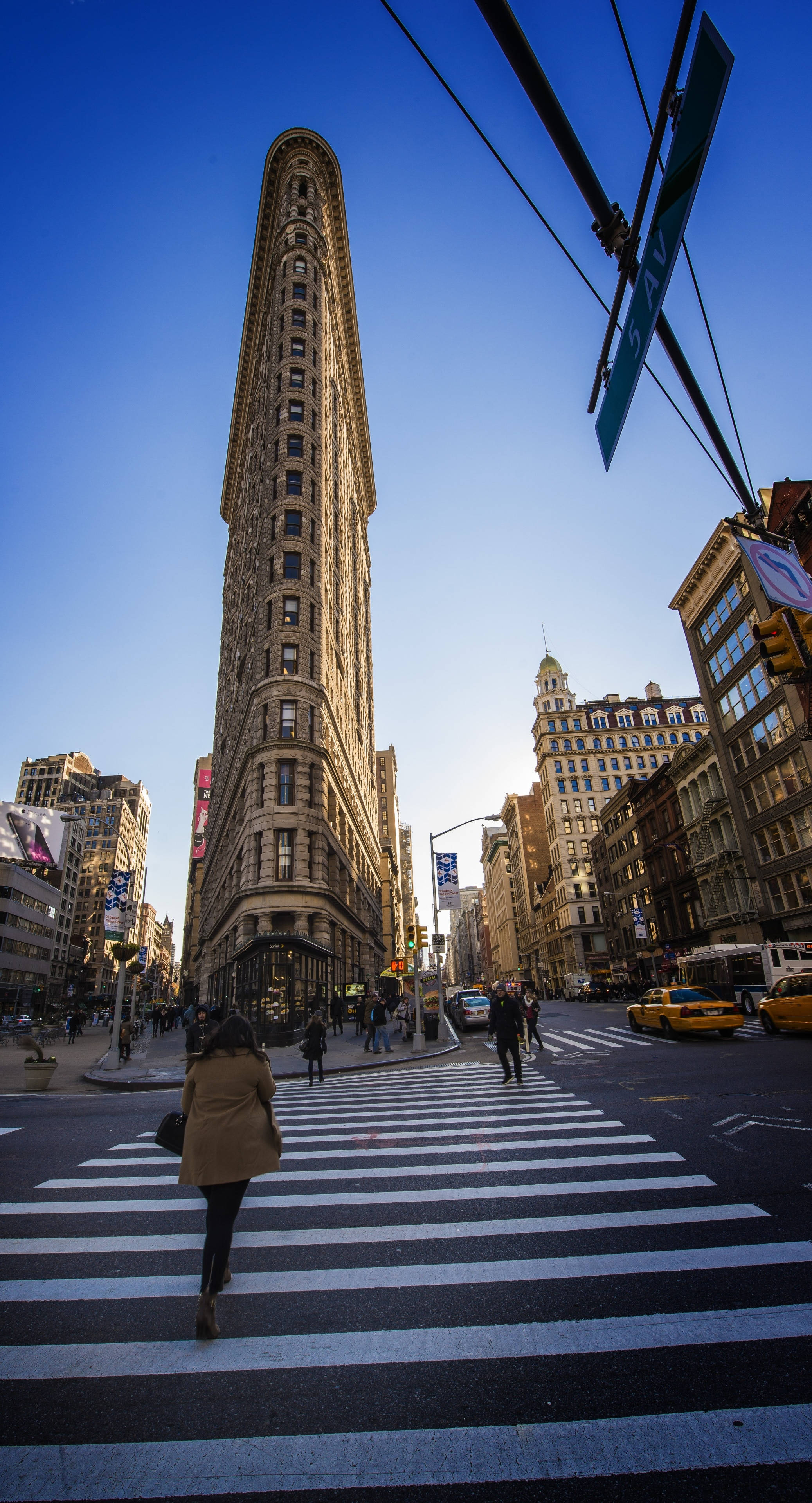 Flatiron Building 桌布 壁紙 — 低角度拍攝捕捉到標誌性的熨斗大廈聳立在繁忙的紐約市街道上，行人在晴朗的藍天下穿過斑馬線。