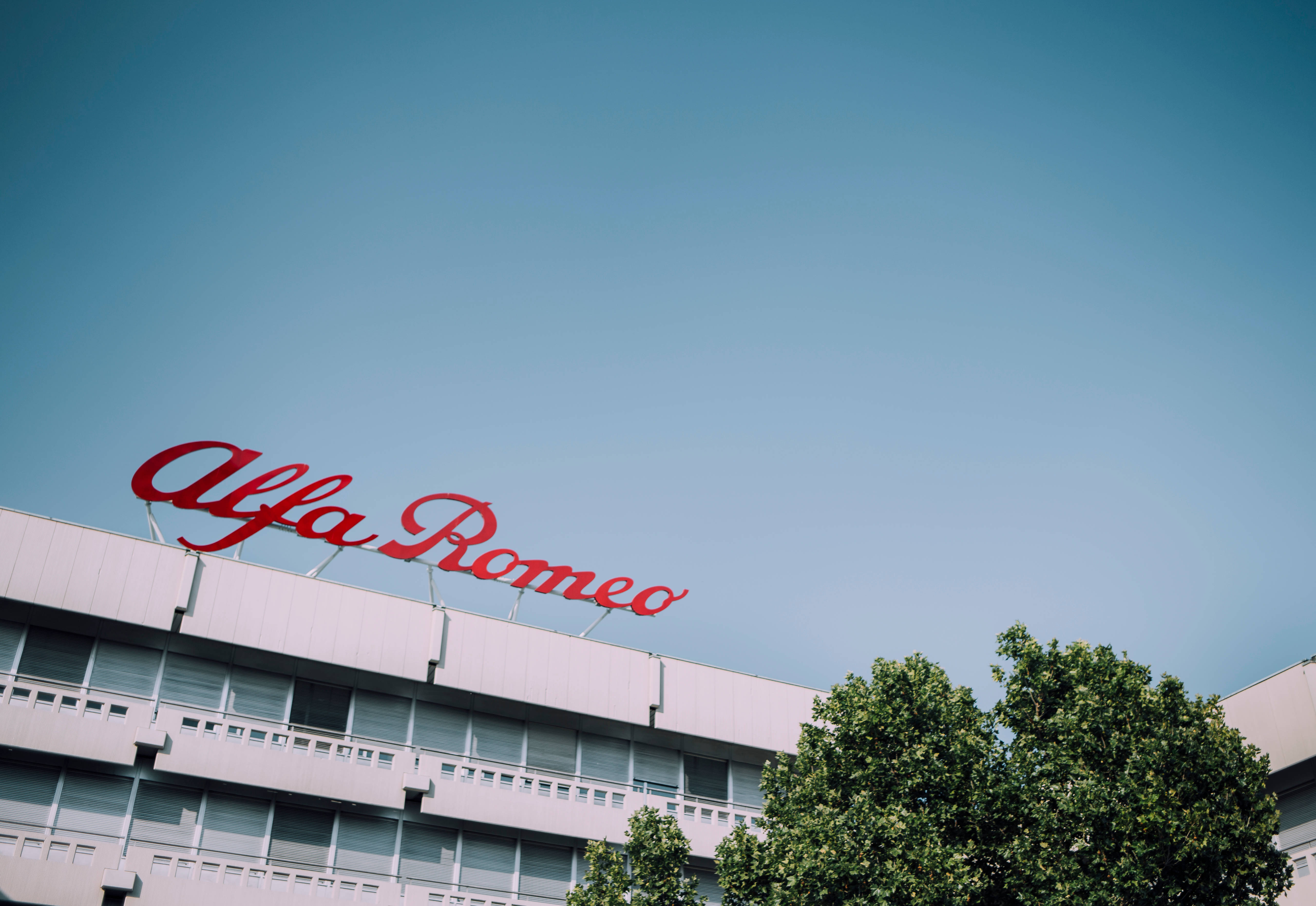 Alfa Romeo 桌布 壁紙 — 低角度拍攝現代建築，頂部有醒目的紅色 Alfa Romeo 標誌，襯托著晴朗的藍天和角落的綠樹。