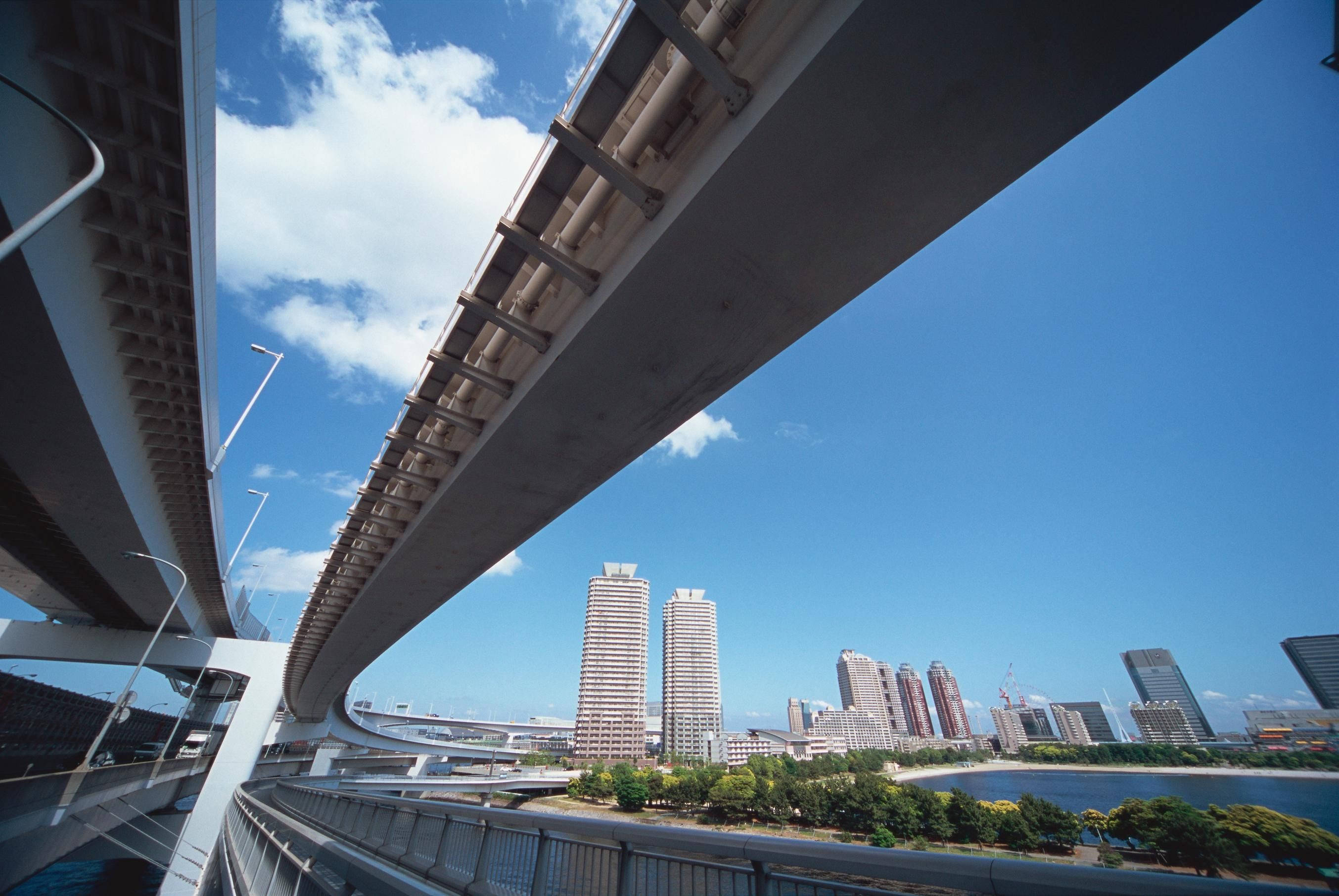 Road 桌布 壁紙 — 低角度拍攝現代混凝土橋樑蜿蜒於河流之上，城市高樓大廈聳立，明亮藍天中飄浮著蓬鬆白雲。