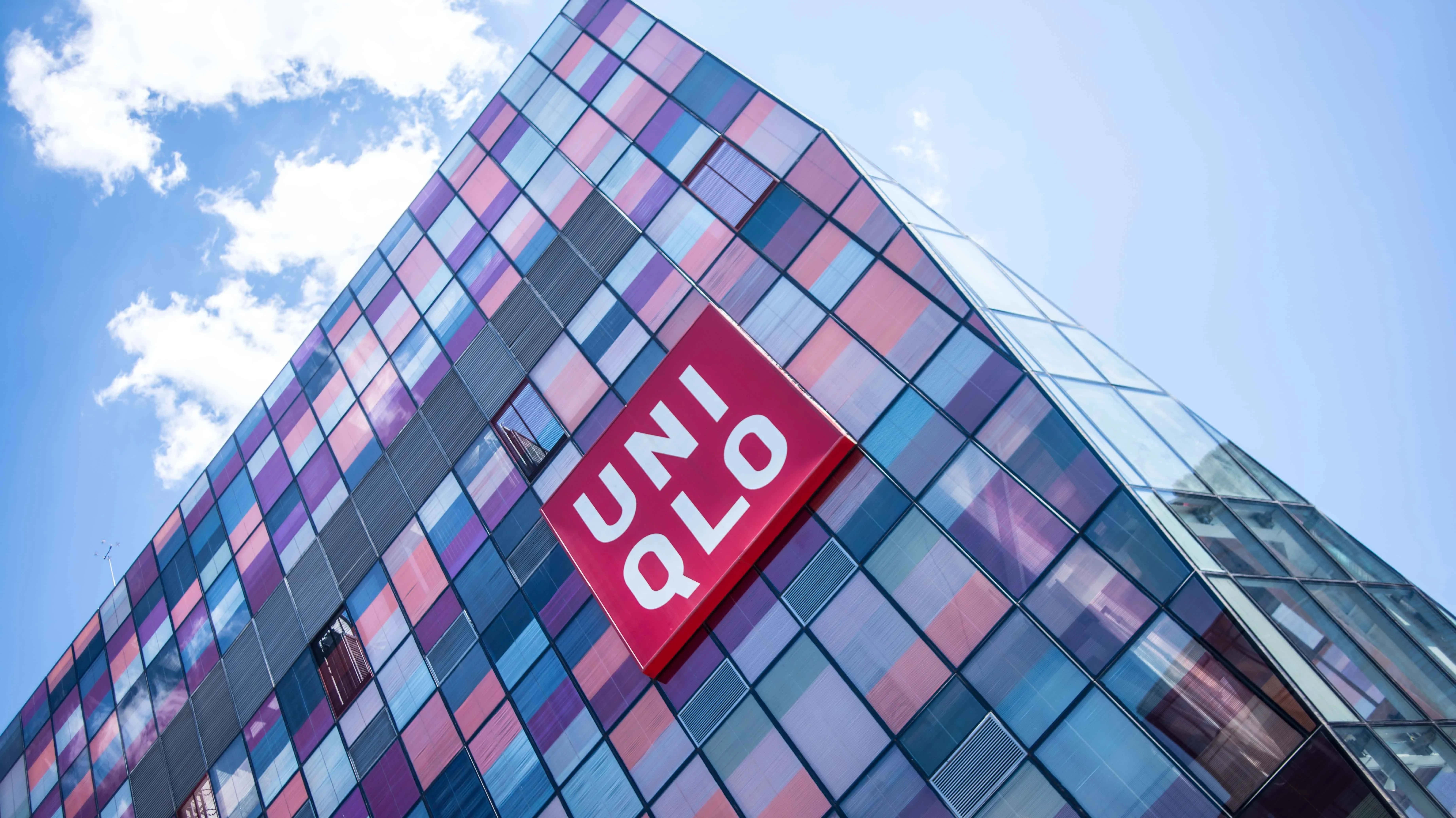 Uniqlo 桌布 壁紙 — 低角度拍攝現代玻璃建築，帶有彩色反光面板和大型紅色UNIQLO標誌，背景是明亮的藍天白雲。