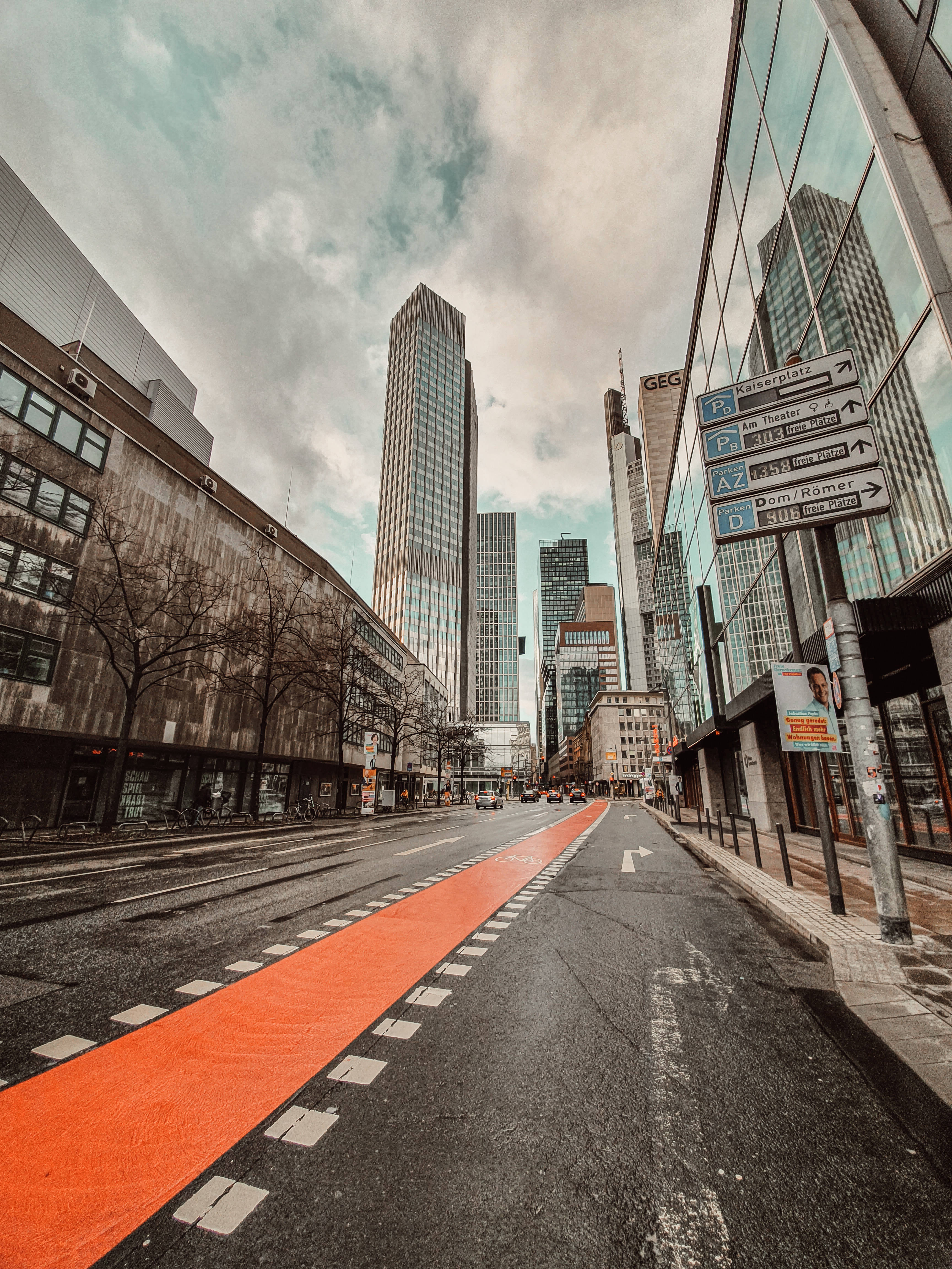 City Street 桌布 壁紙 — 低角度拍攝的城市街道，一條大膽的橙色自行車道通向雲層下高聳的玻璃摩天大樓。