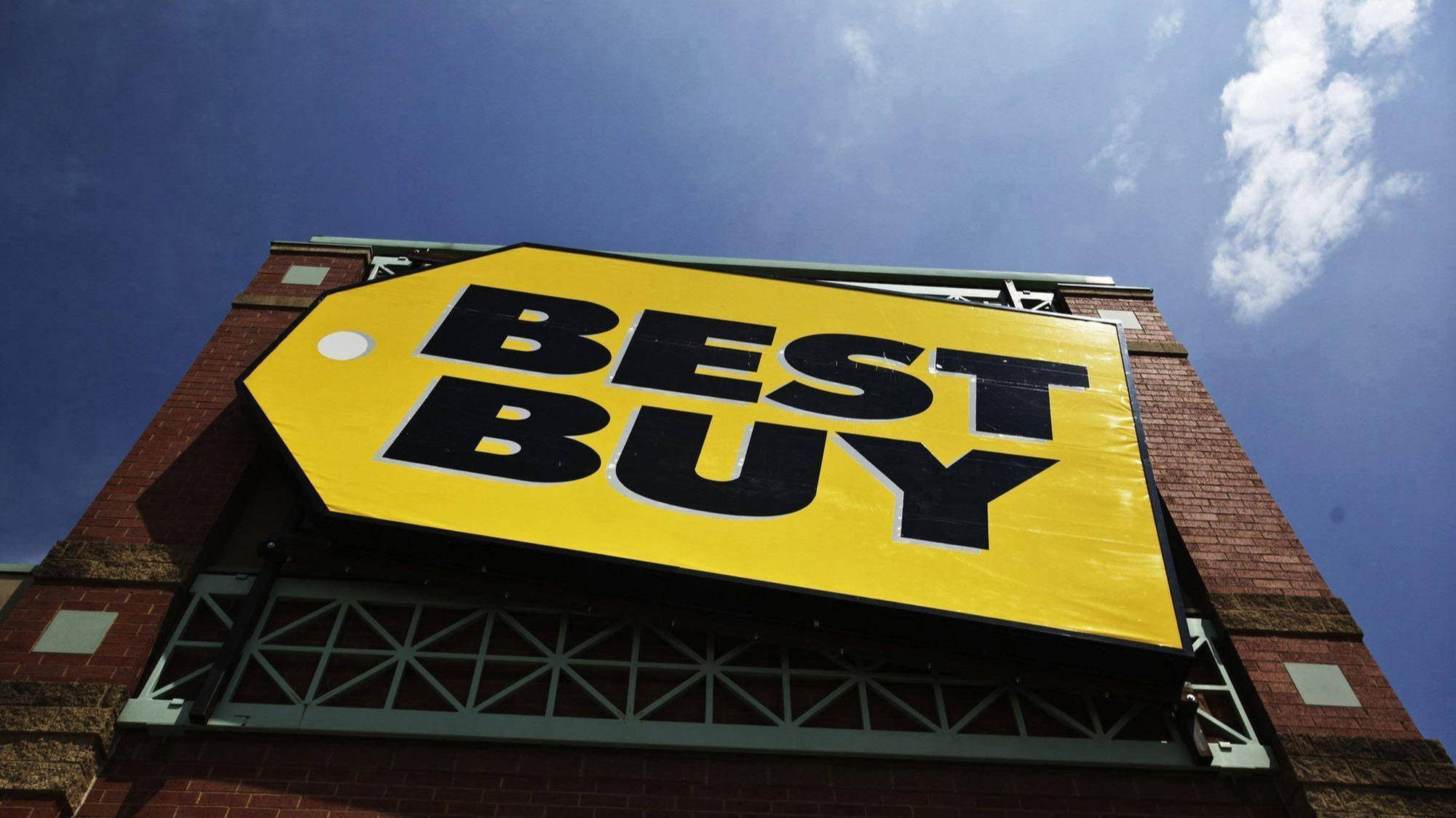 buy 桌布 壁紙 — 低角度拍攝的明亮黃色Best Buy標誌，安裝在磚砌建築上，背景是清澈藍天和零星白雲。