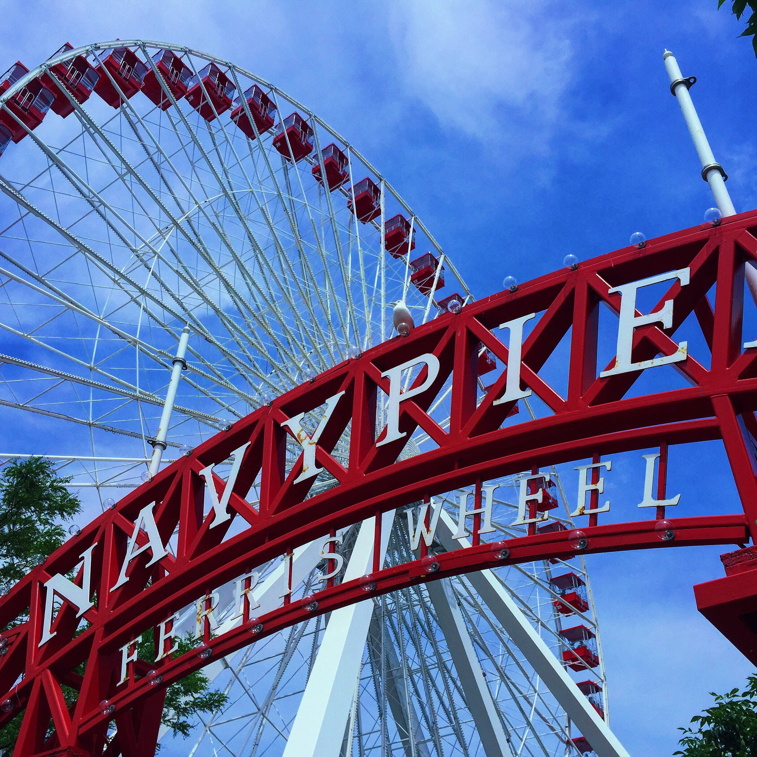 雲朵 桌布 壁紙 — 低角度拍攝的紅色拱門，上面有「NAVYPIE FERRIS WHEEL」字樣，背景是明亮的藍天和一個大型白色摩天輪。