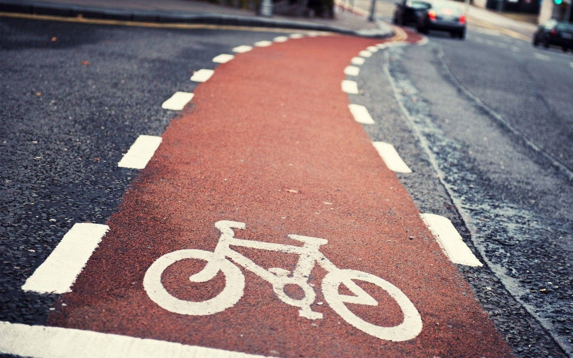 Bicycle 桌布 壁紙 — 低角度拍攝的紅色自行車道，帶有白色自行車標誌，蜿蜒穿過城市街道。