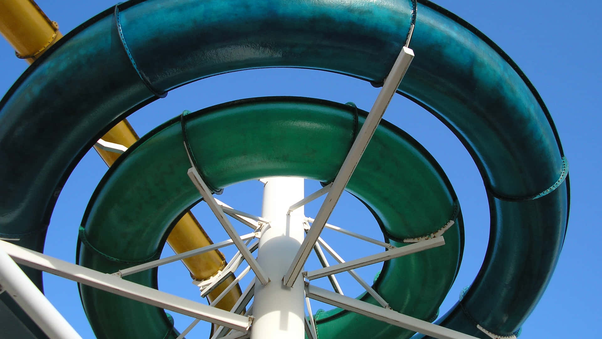 Swimming Pool Slide. 壁紙 — 低角度拍攝的螺旋綠色水上滑梯，以清澈藍天和白色金屬支架為背景。