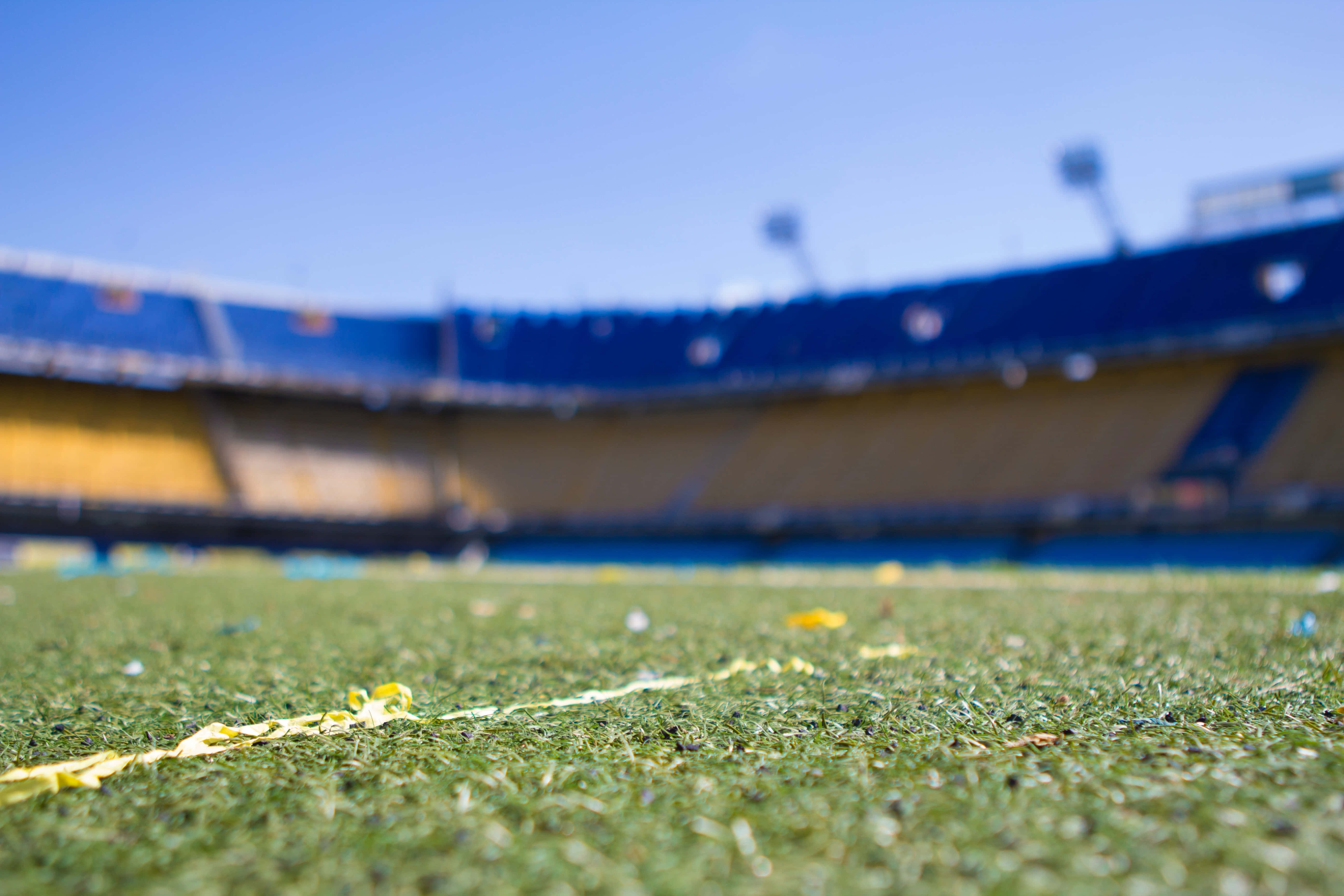 Stadium 桌布 壁紙 — 低角度拍攝綠色足球場草地，黃色紙屑散落，背景是明亮的藍天和模糊的體育館看台。