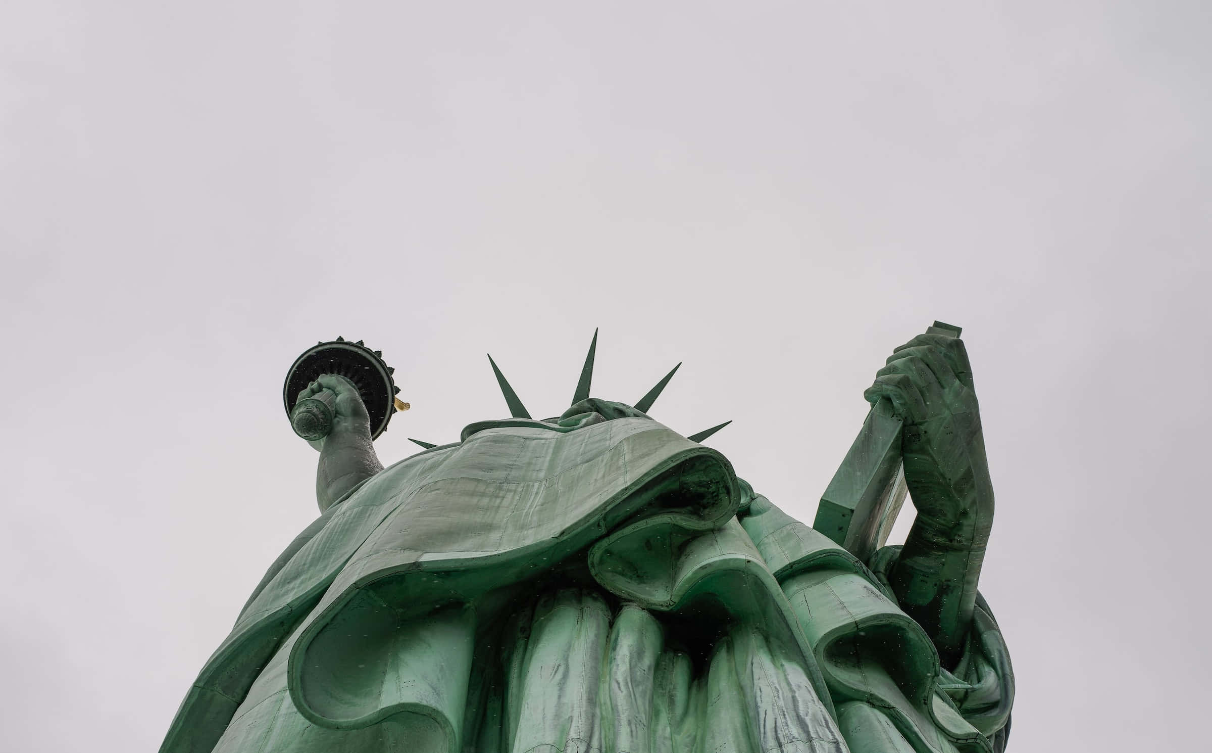 New York 桌布 壁紙 — 低角度拍攝綠色銅製自由女神像手持火炬和石碑，背景為灰色天空。