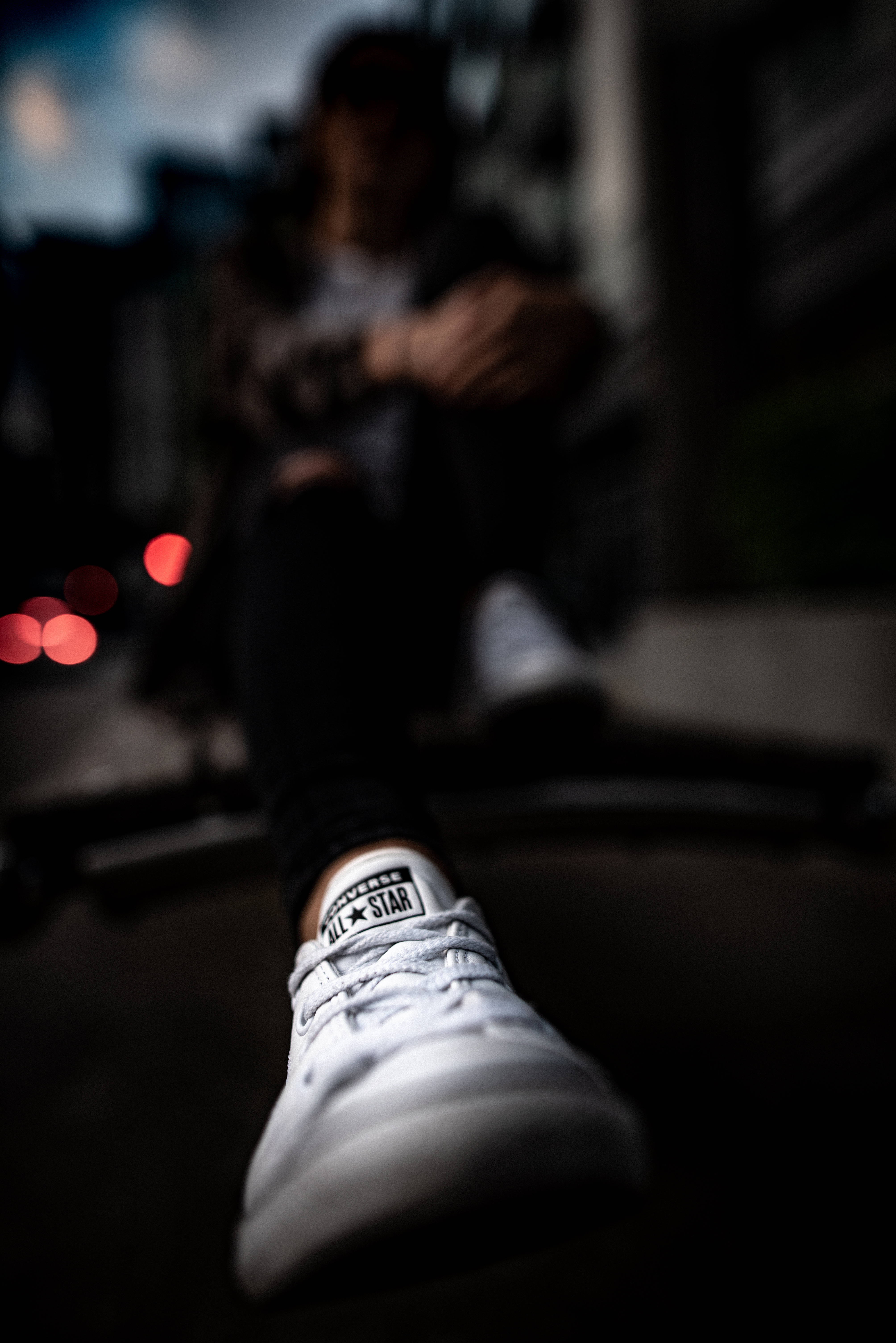 Converse 桌布 壁紙 — 低角度拍攝聚焦於一雙白色Converse運動鞋，背景是模糊的人坐在昏暗的城市環境中。