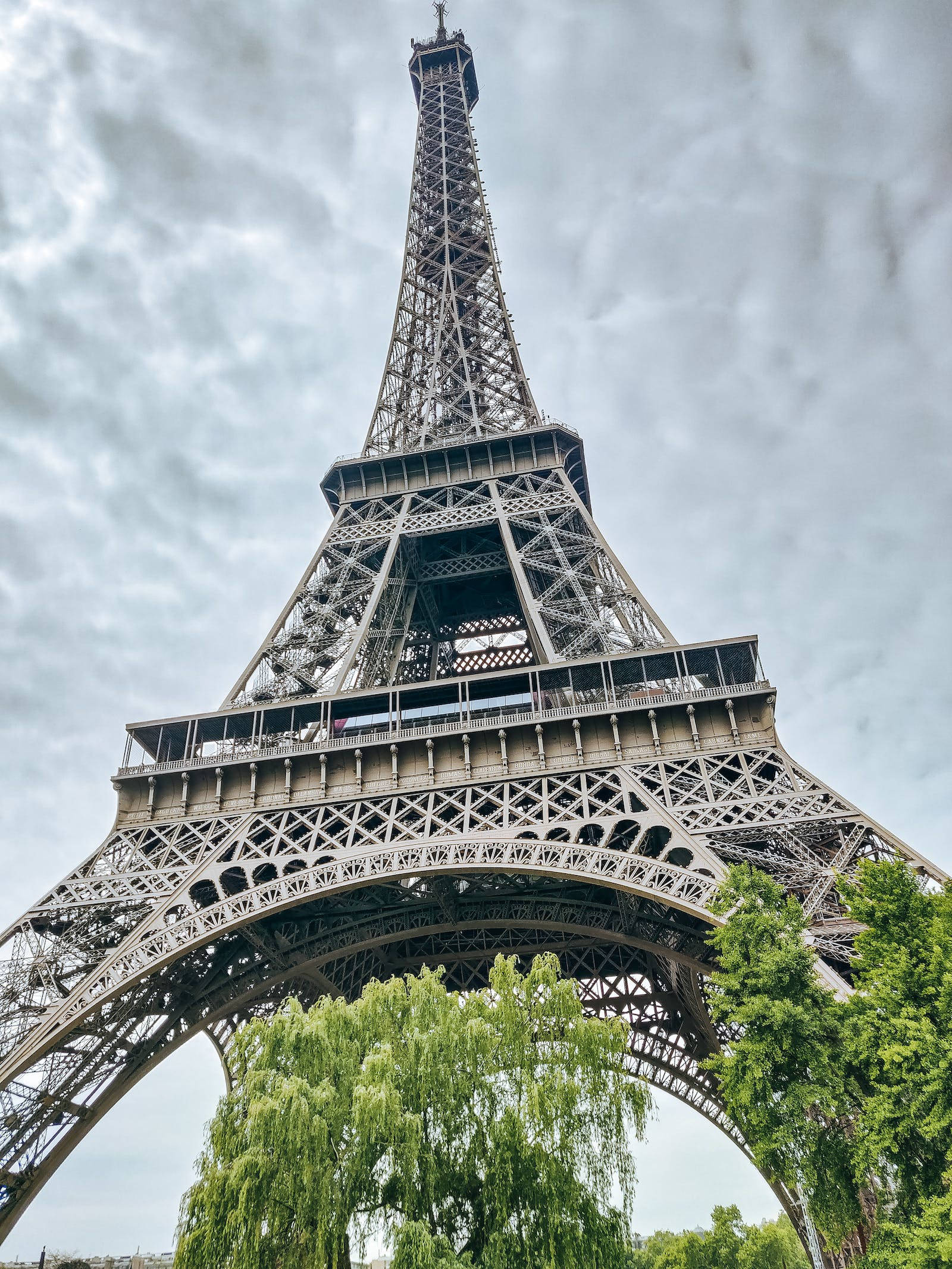 france iPhone 桌布 壁紙 — 低角度拍攝艾菲爾鐵塔複雜的鐵製格子結構，背景為多雲的天空，底部有茂密的綠樹。