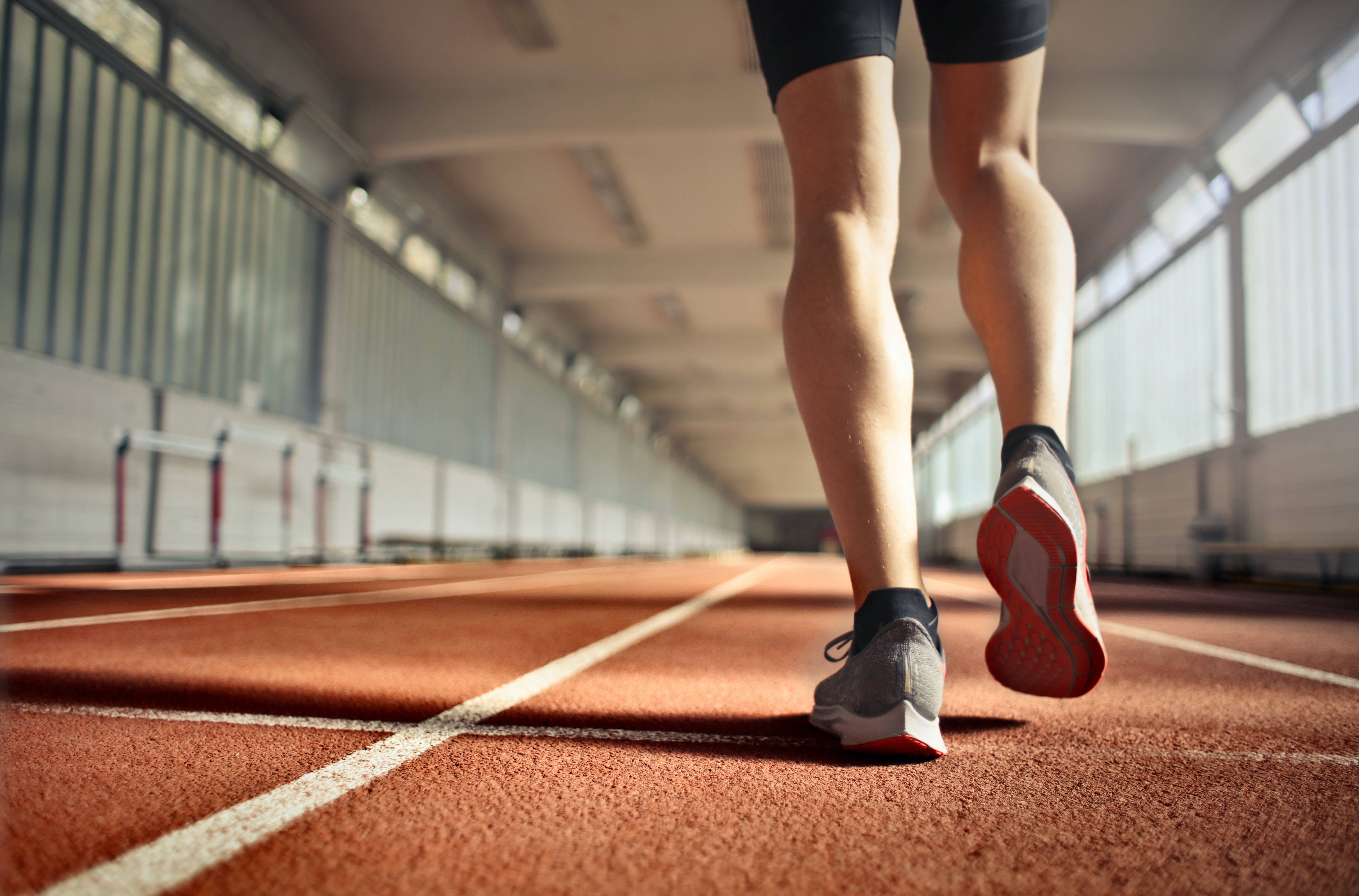 Fitness 桌布 壁紙 — 低角度拍攝跑者雙腿在紅色室內跑道衝刺，跑道有白色線條，背景有模糊的跨欄。