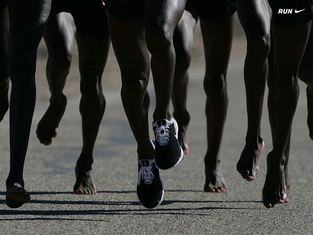 Run 桌布 壁紙 — 低角度拍攝跑者雙腿在運動中的畫面，一人穿著運動鞋，其他赤腳在陽光普照的柏油路上。