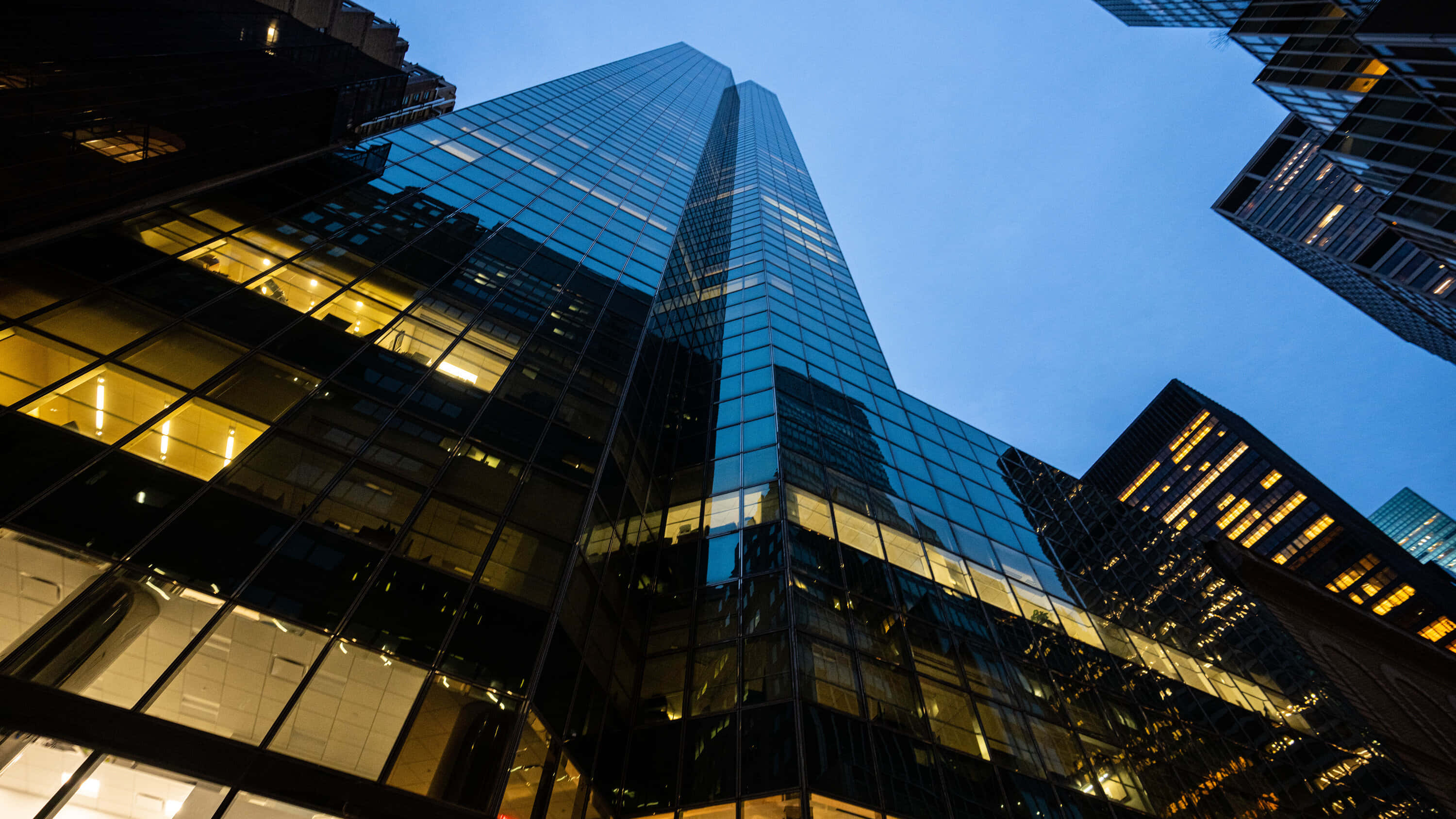 Wall Street 桌布 壁紙 — 低角度拍攝高聳的玻璃摩天大樓刺穿暮色天空，溫暖的室內燈光在冷藍色調中閃耀。
