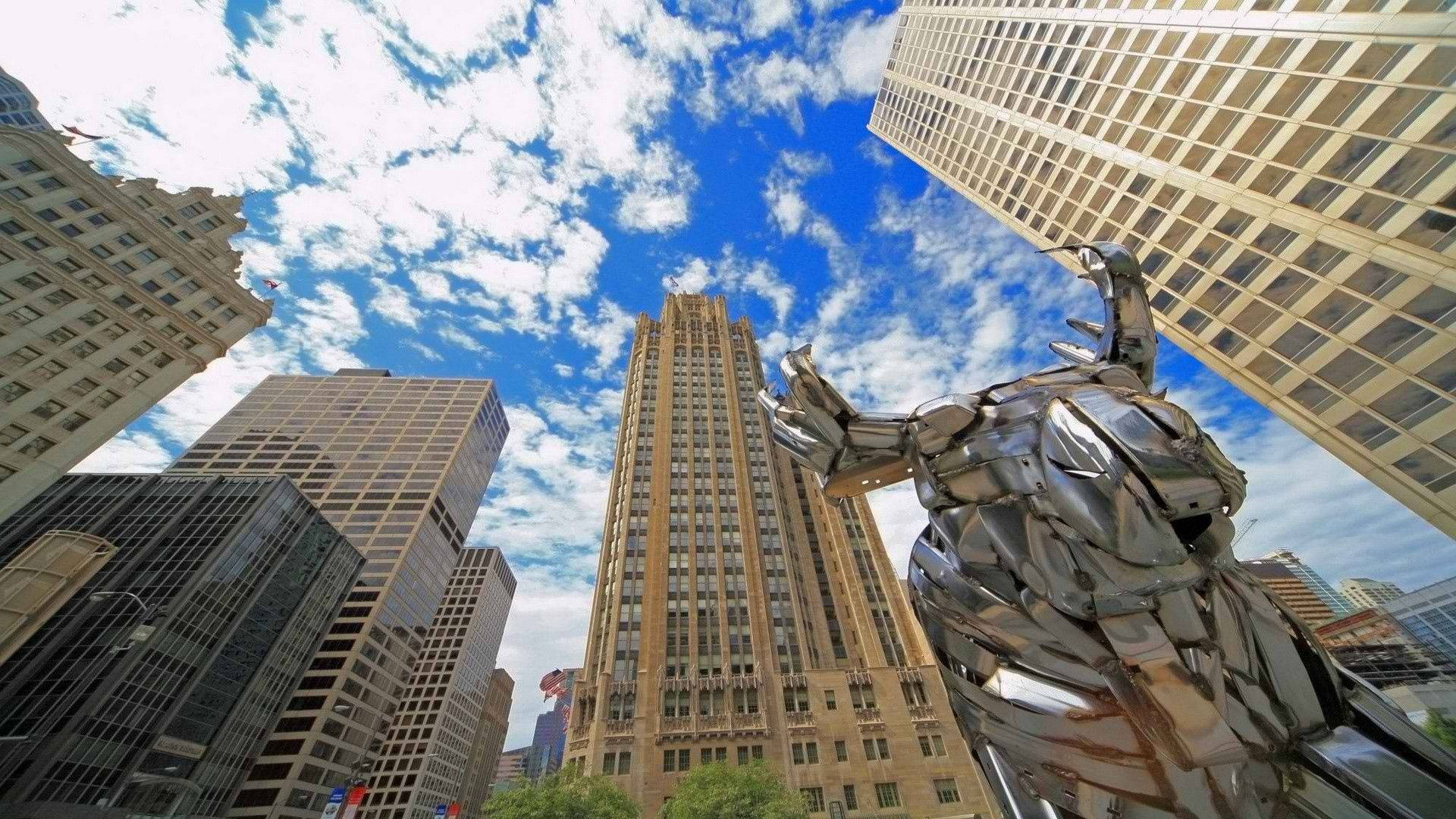 芝加哥 桌布 壁紙 — 低角度視角捕捉到一座反射性金屬雕塑，聳立在摩天大樓之間，天空湛藍，雲朵點綴。