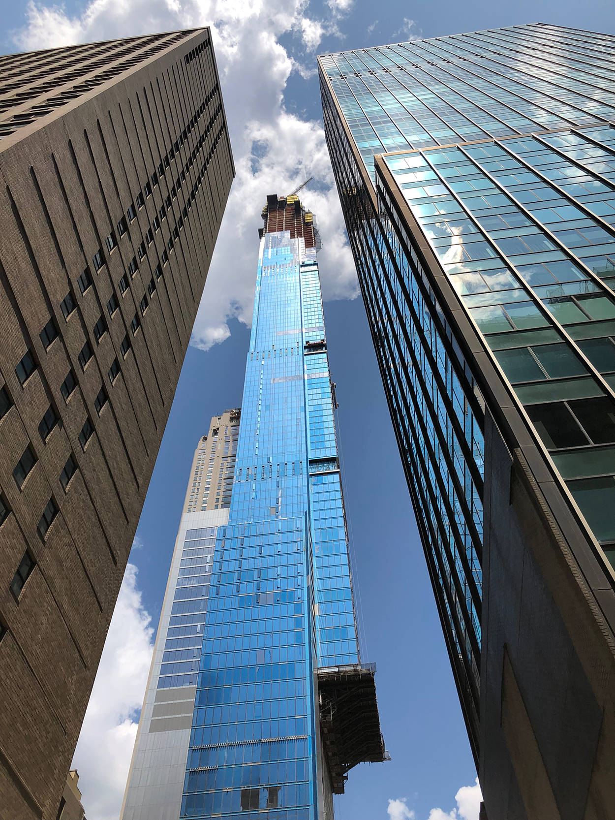 Buildings 桌布 壁紙 — 低角度視角捕捉到一座高聳的藍色玻璃摩天大樓正在兩棟舊建築之間施工，背景是明亮的藍天和蓬鬆的白雲。