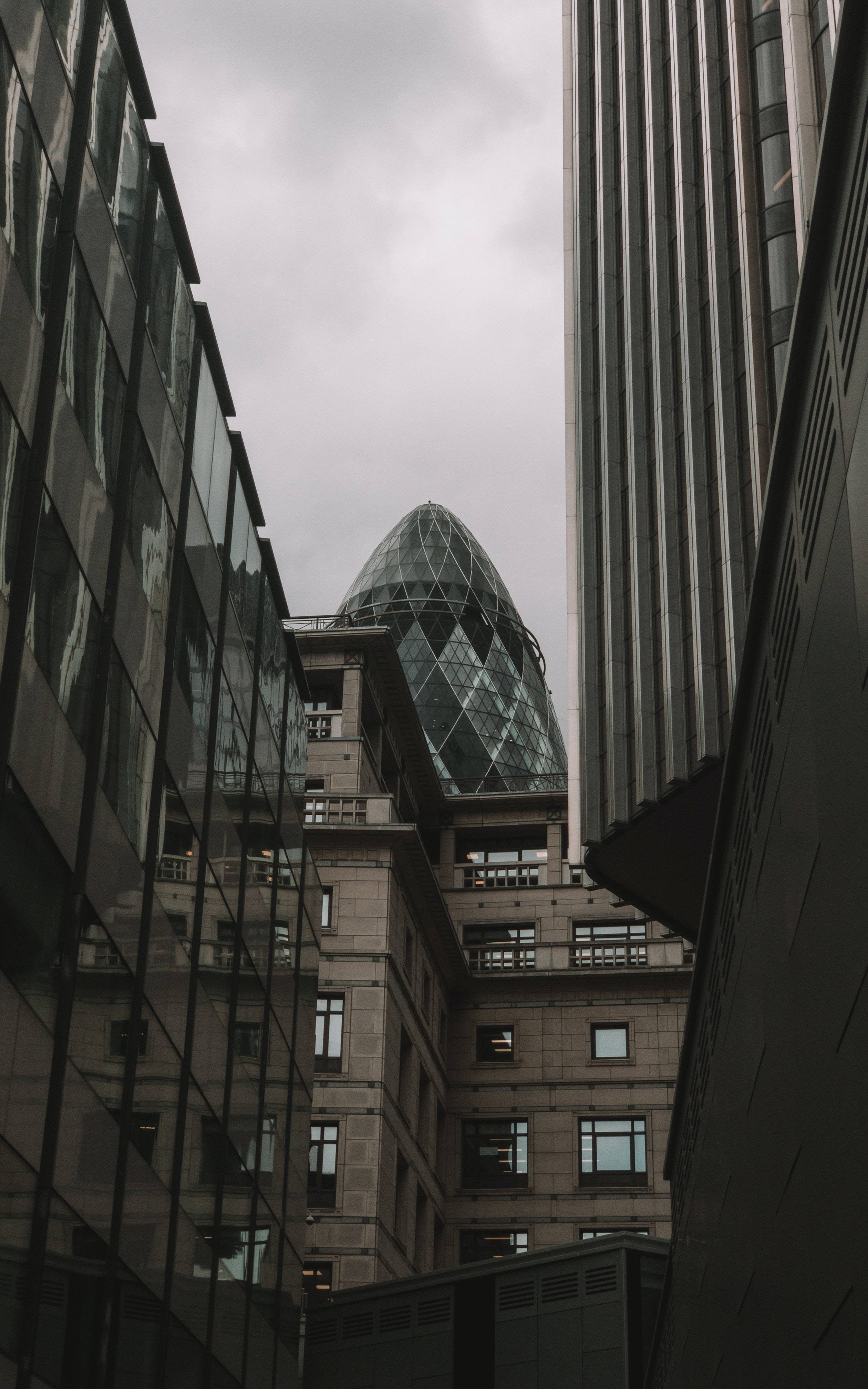 Buildings 桌布 壁紙 — 低角度觀看Gherkin摩天大樓在狹窄城市建築之間窺視，天空多雲。
