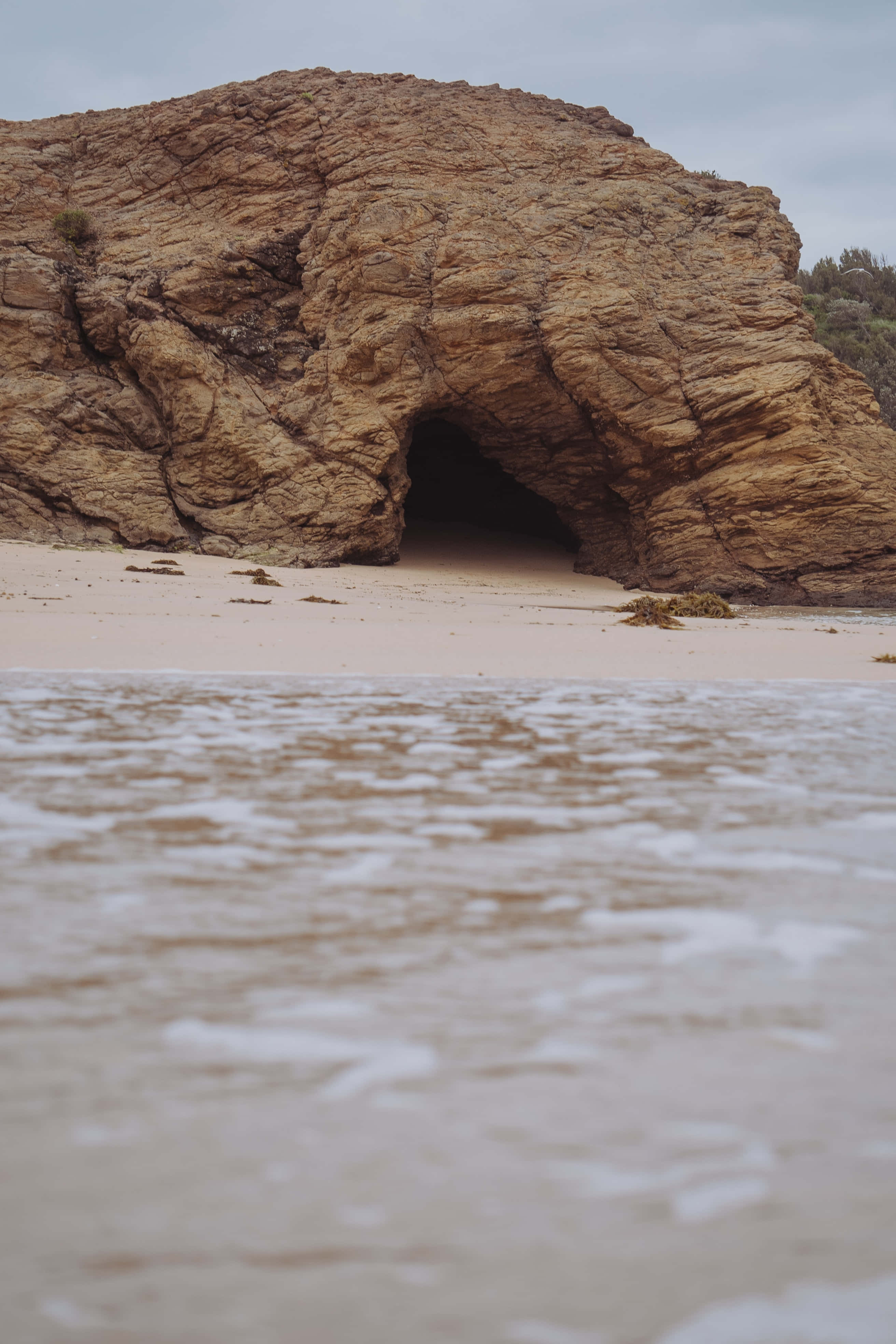 Cave 桌布 壁紙 — 低角度觀看溫柔的海浪拍打沙灘，前方是巨大的岩石拱門，天空多雲。