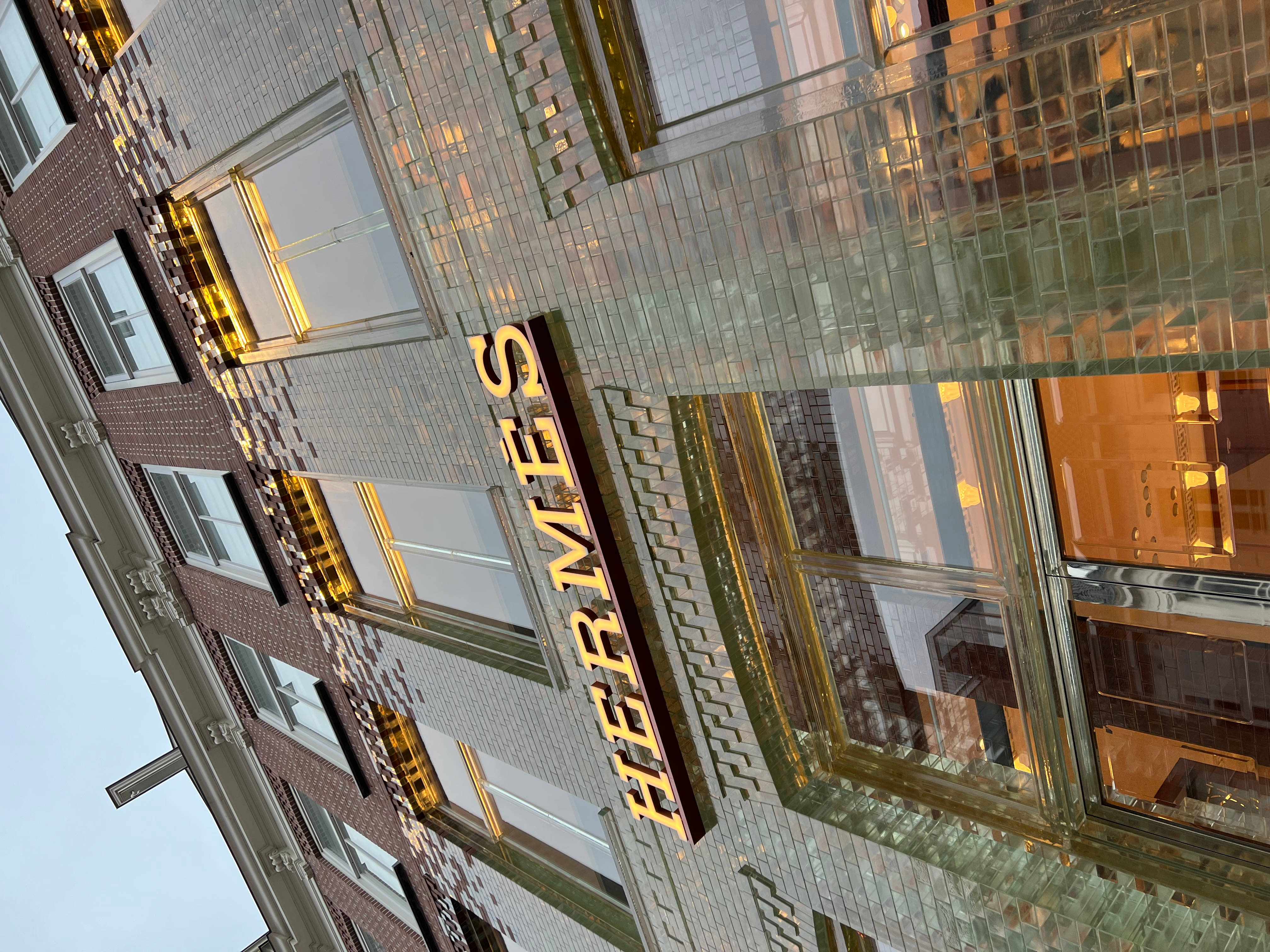 Hermes 桌布 壁紙 — 低角度觀看豪華愛馬仕商店，金色字體和玻璃外牆反射溫暖的室內燈光。