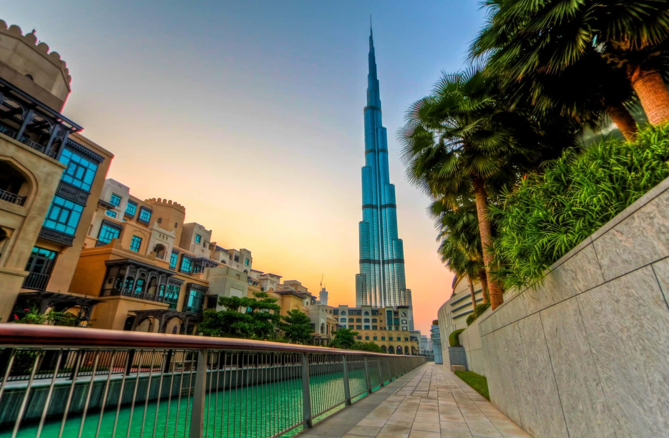 Burj Khalifa 桌布 壁紙 — 低角度觀賞哈里發塔在日落時分聳立於運河步道上，附近有棕櫚樹和傳統建築。