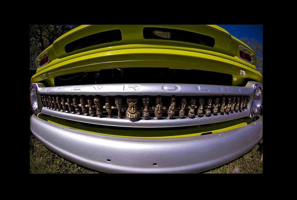 Car Grill 桌布 壁紙 — 低角度魚眼鏡頭拍攝捕捉到一輛復古萊姆綠色雪佛蘭卡車的鍍鉻格柵，裝飾著部落面具，背景是晴朗的藍天。