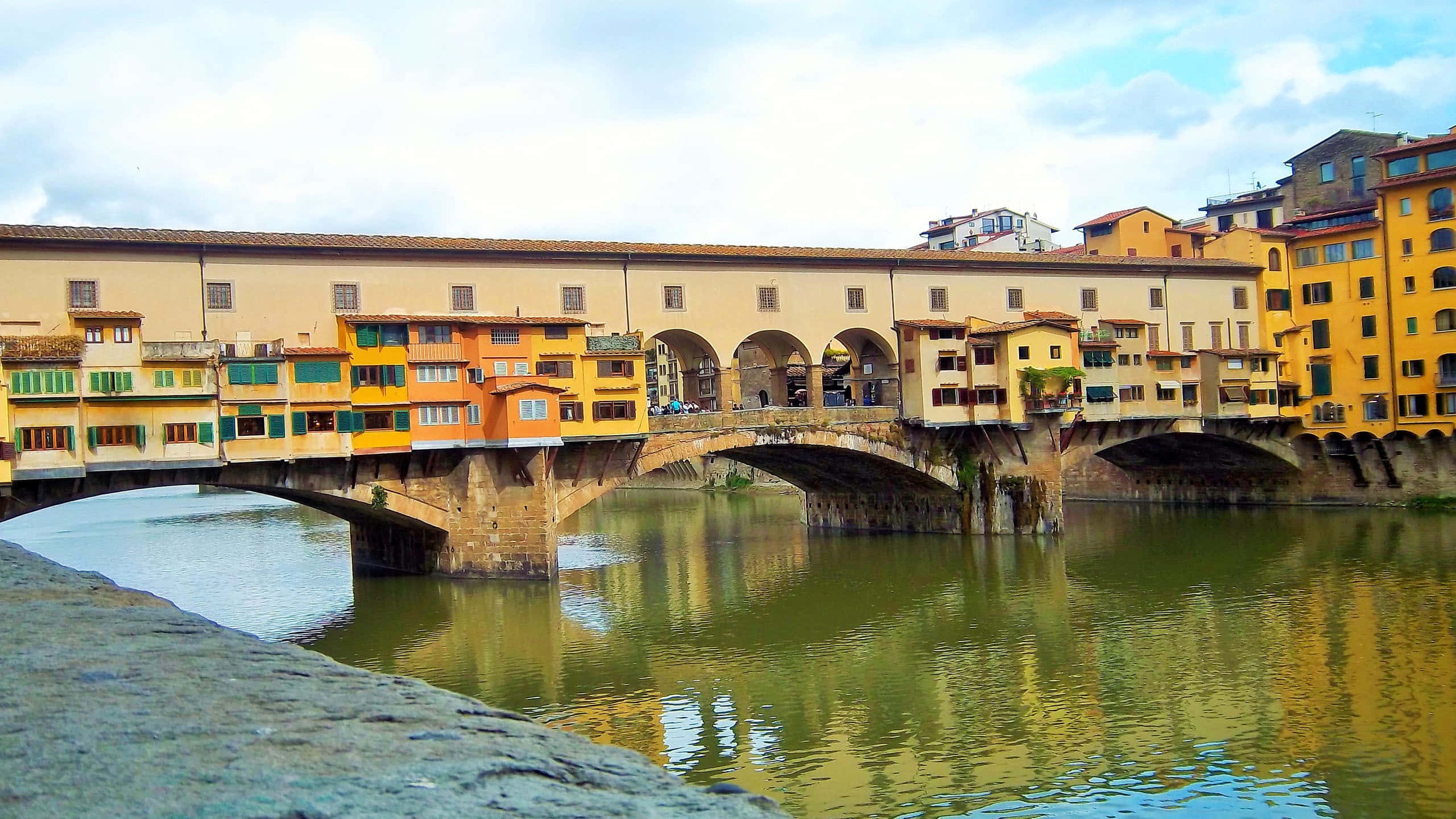 水 桌布 壁紙 — 佛羅倫斯的舊橋（Ponte Vecchio）拱跨阿諾河，石造結構兩旁排列著色彩繽紛的商店，天空部分多雲。
