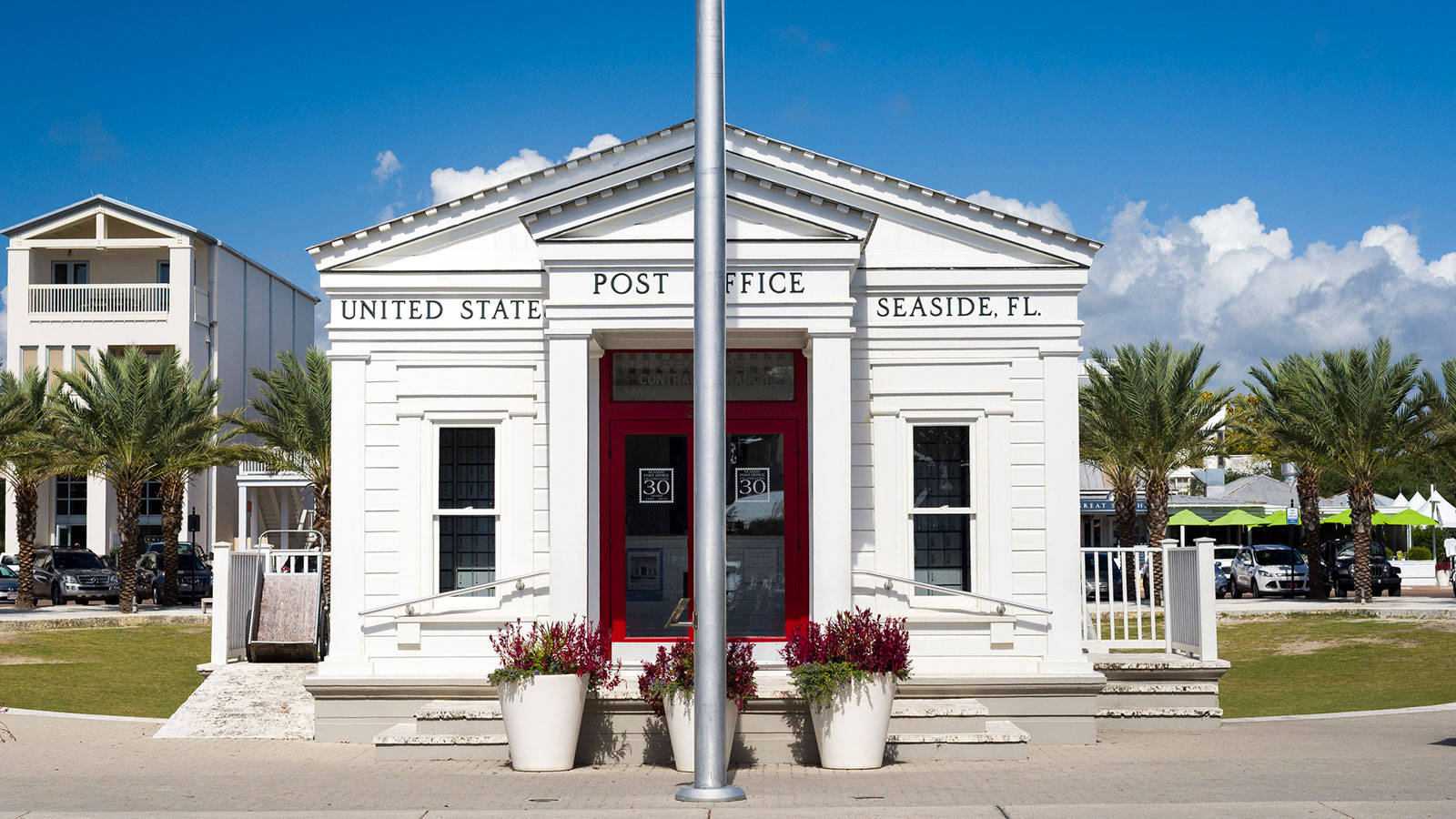 Post Office 桌布 壁紙 — 佛羅里達州海邊小鎮 Seaside 的一座明亮白色古典郵局，有紅色門和棕櫚樹，背景是藍天。