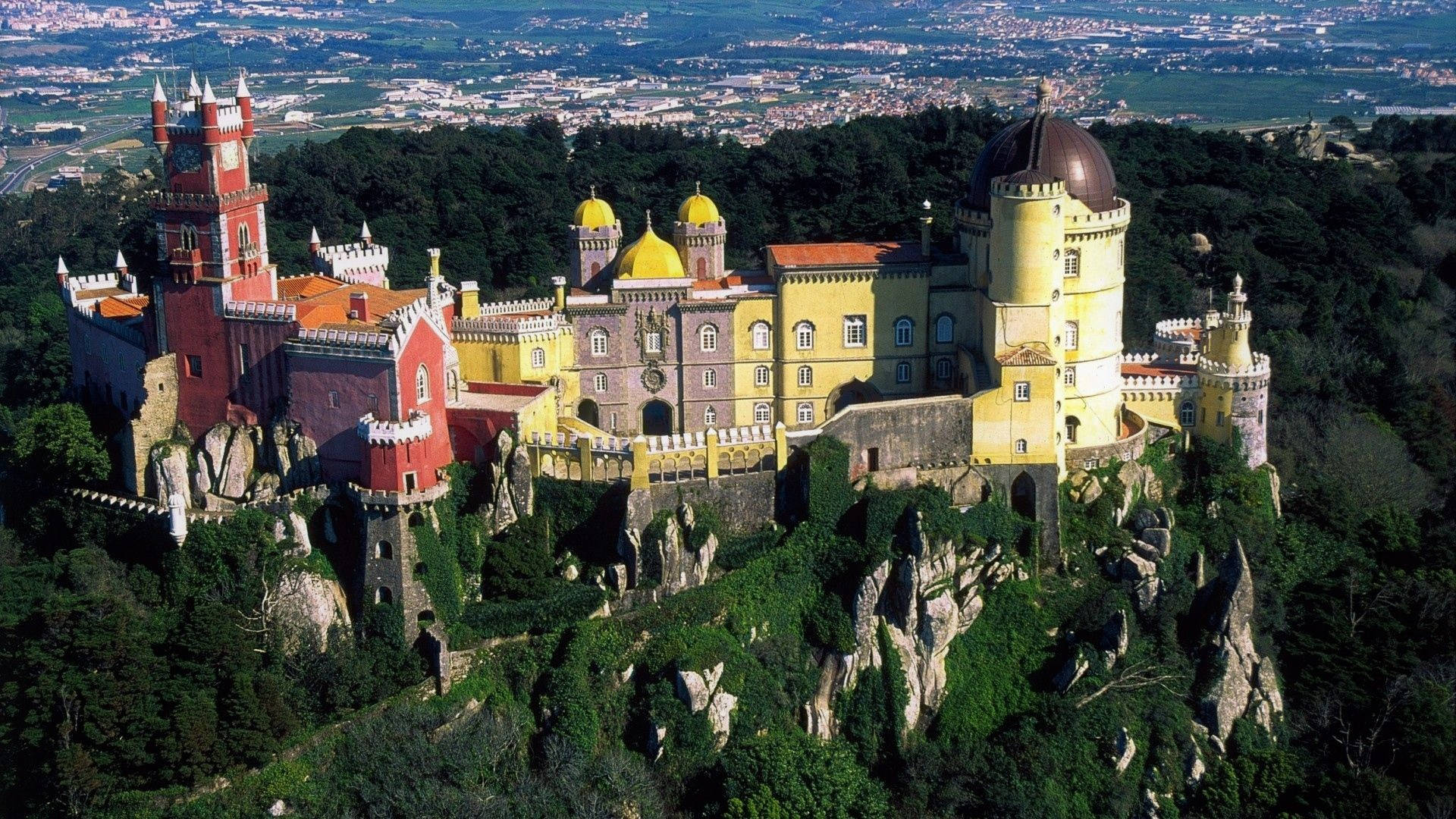 Portugal 桌布 壁紙 — 佩納宮的鳥瞰圖，戲劇性地坐落在岩石山坡上，周圍環繞著茂密的綠色森林，俯瞰著廣闊的山谷城鎮。